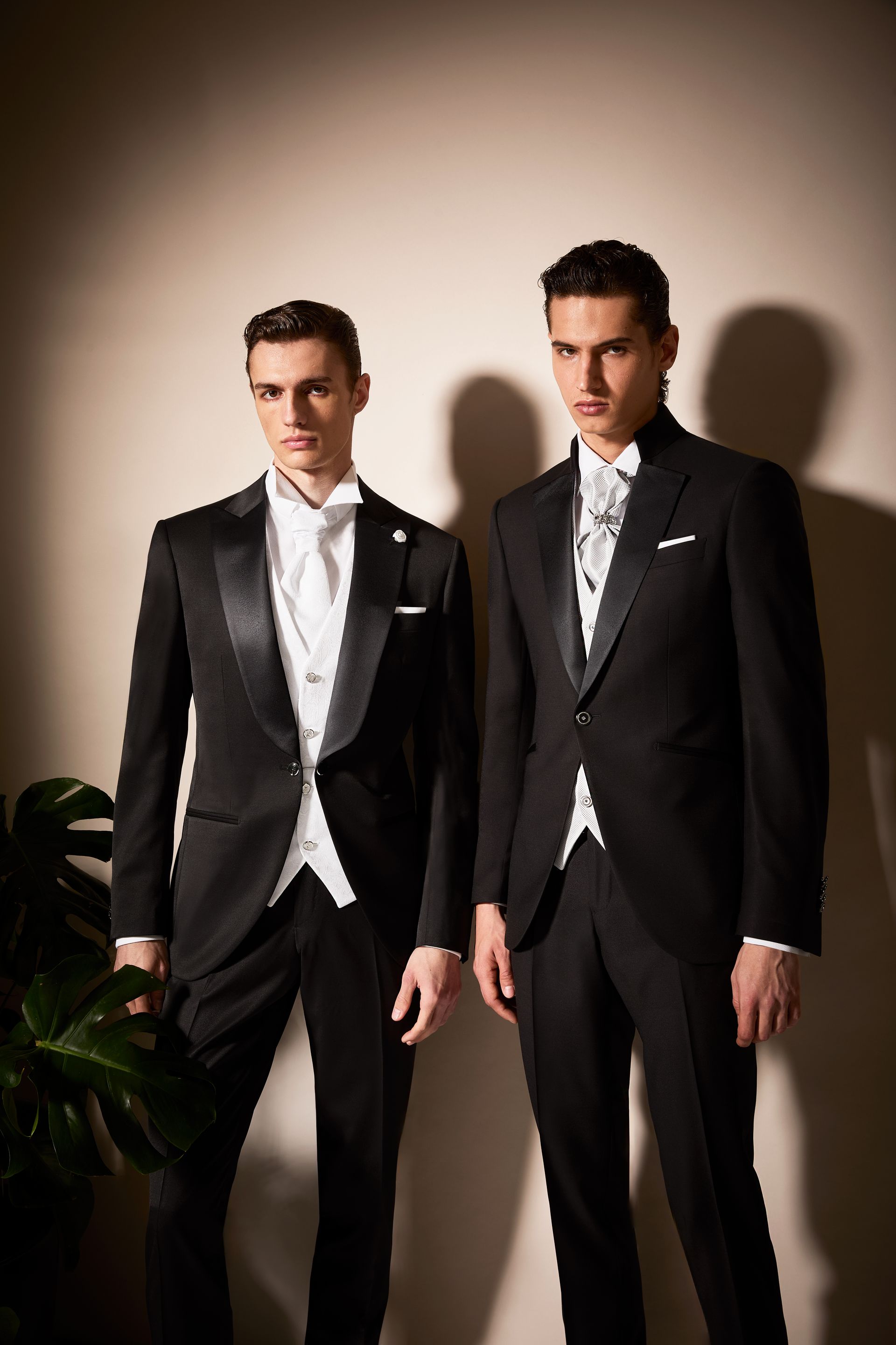 Due uomini in abiti neri formali sono in piedi su uno sfondo neutro. Indossano gilet e cravatta bianchi.
