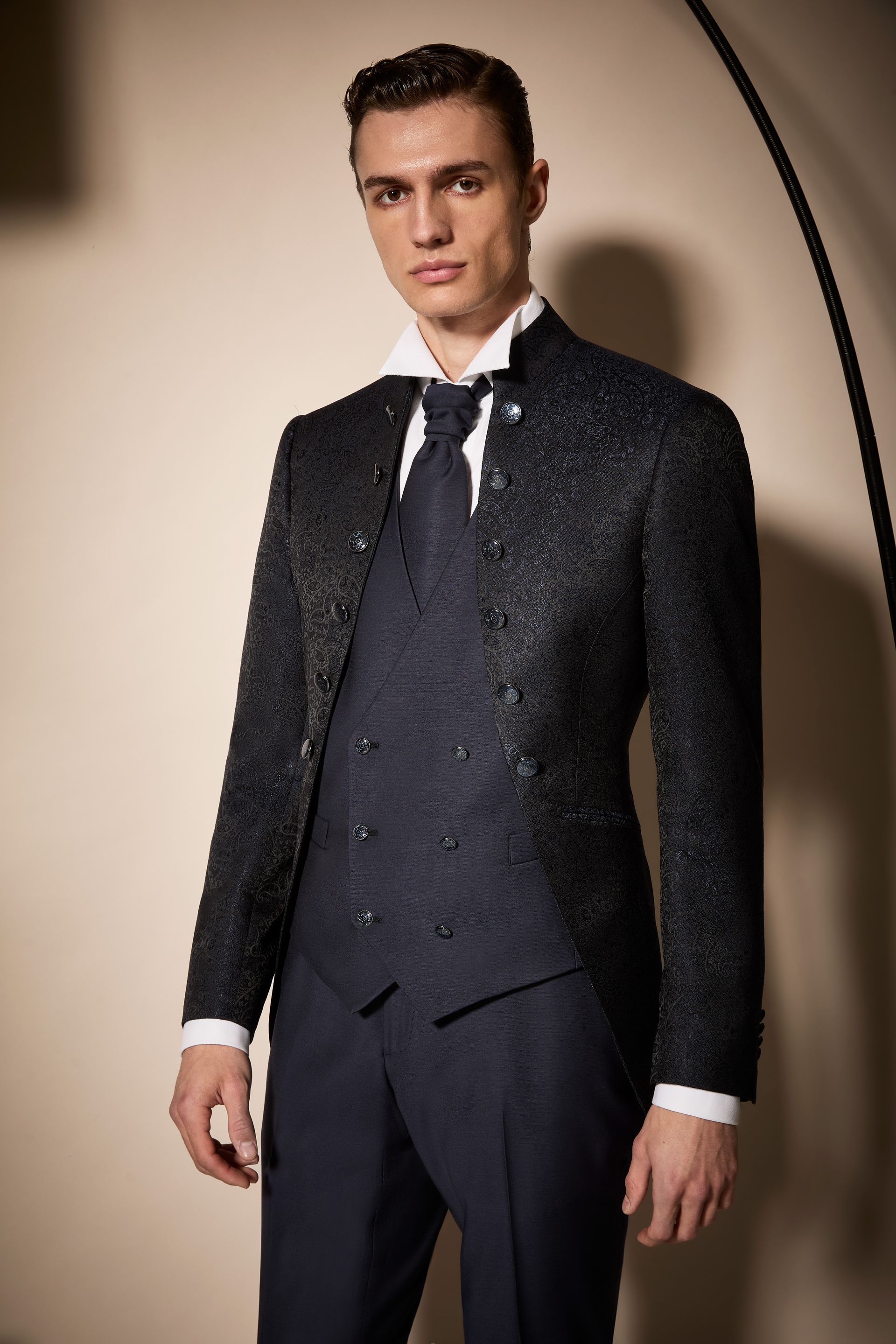 Uomo in abito scuro formale, camicia bianca e cravatta, in posa in studio.