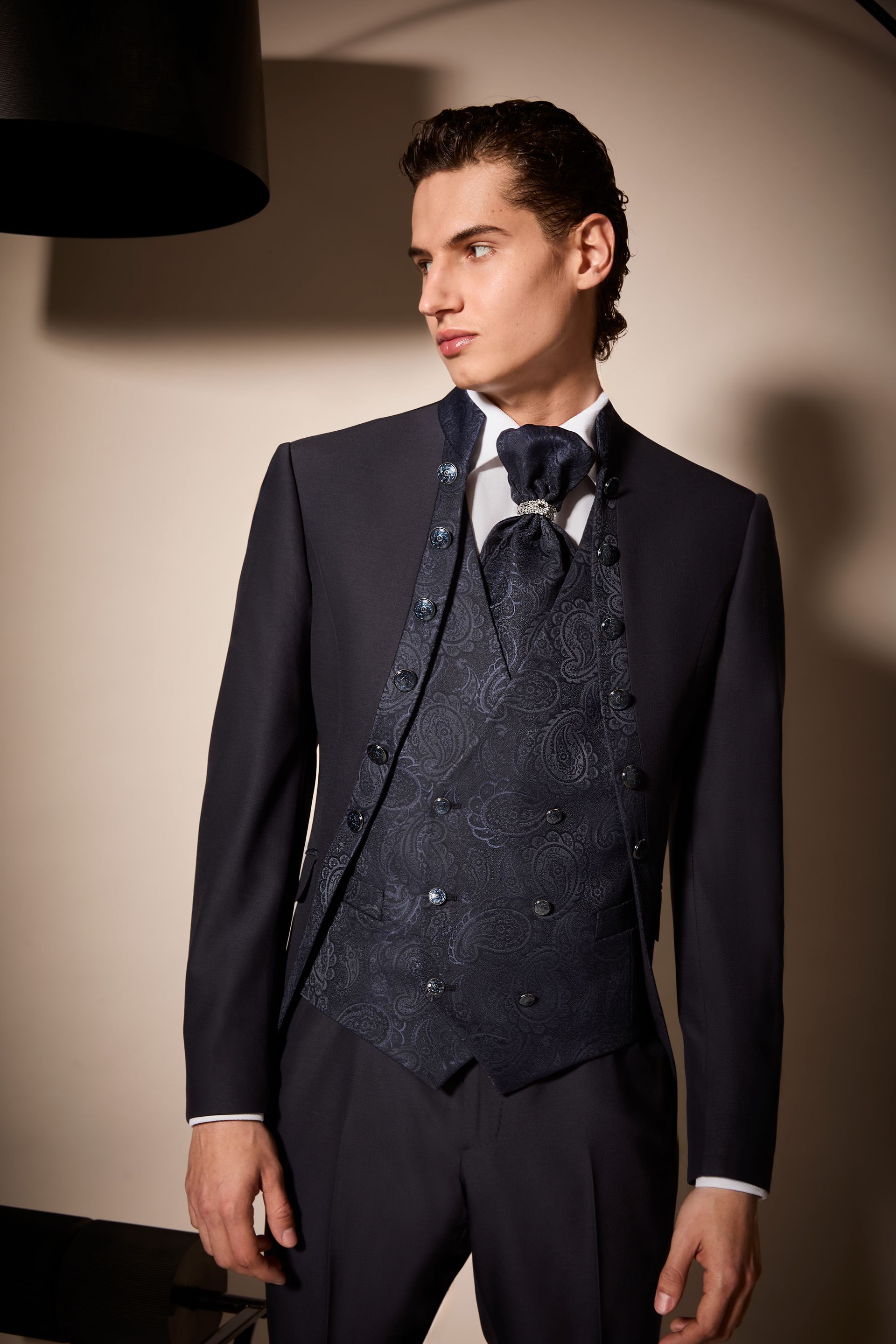 Uomo in abito scuro con gilet fantasia e papillon, in piedi in uno studio fotografico.