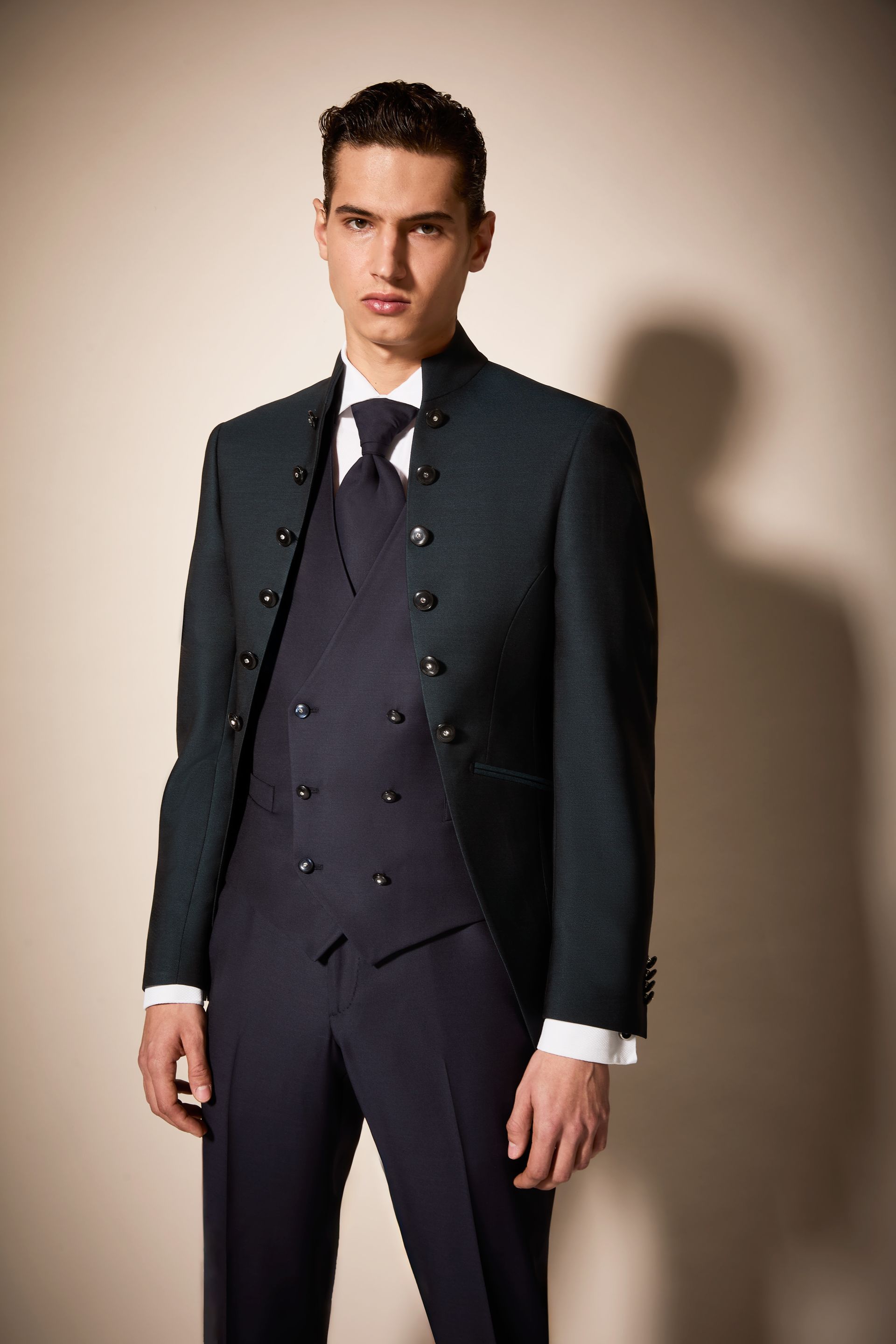 Un uomo in abito verde acqua scuro, in piedi contro un muro beige, guarda avanti.