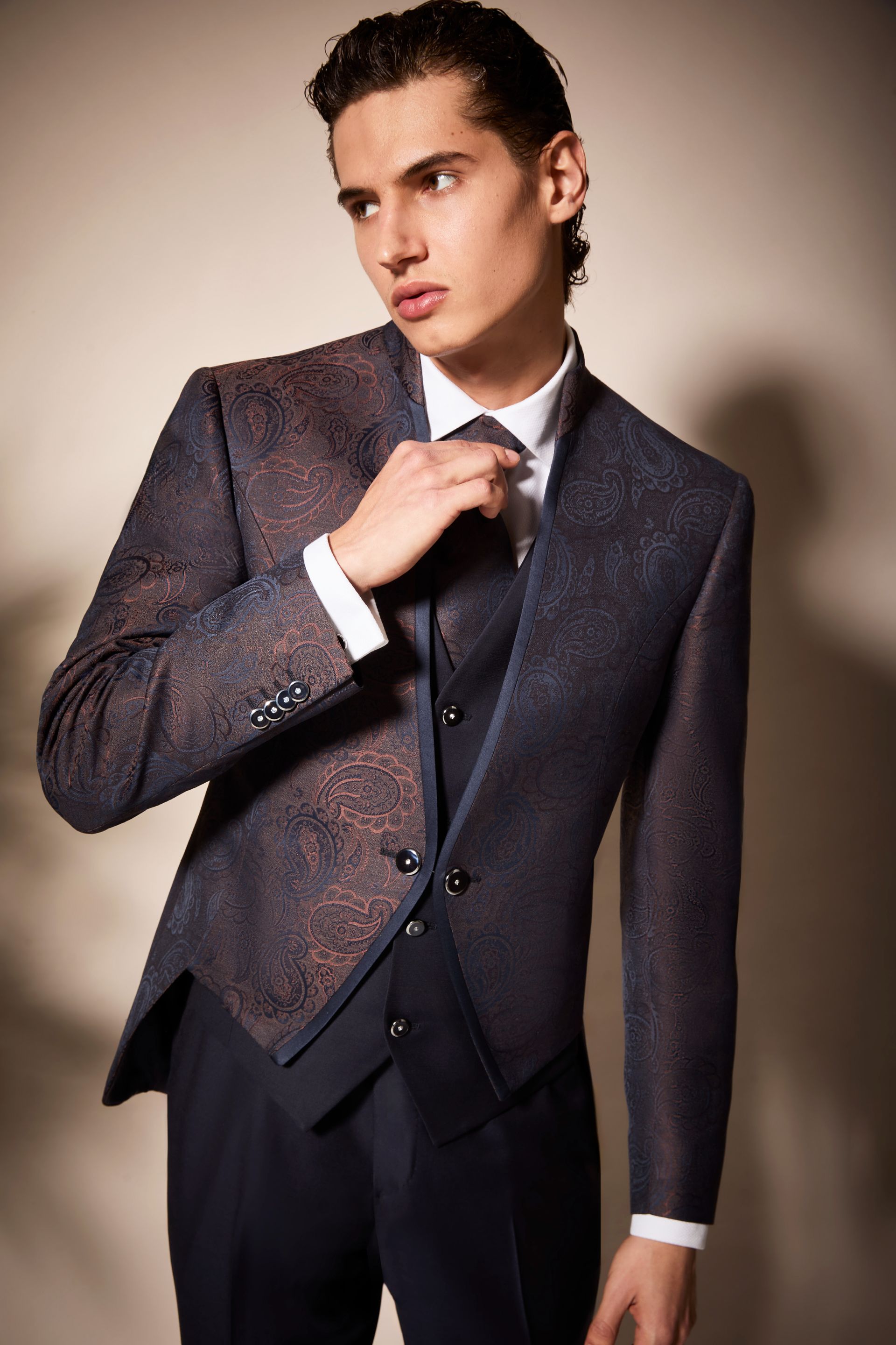 Uomo in abito blu fantasia che sistema la cravatta. Camicia bianca, gilet scuro e sfondo neutro.