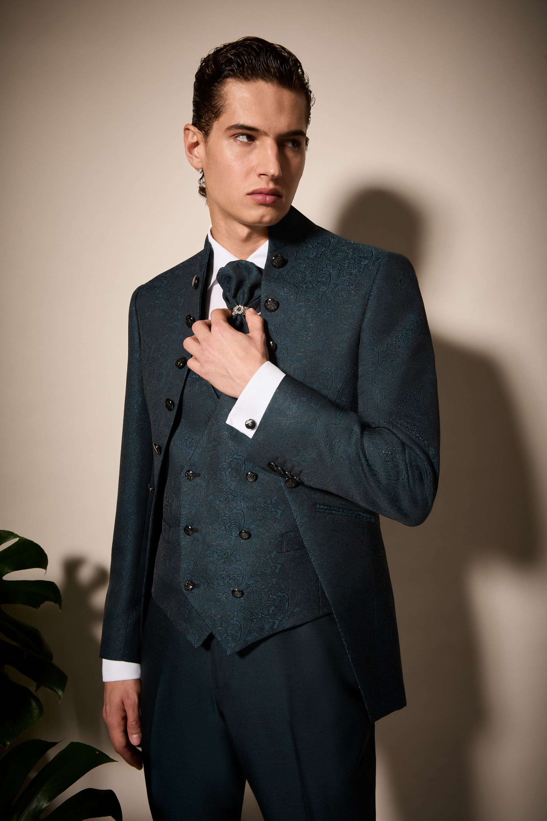 Uomo in abito verde acqua che si sistema la cravatta. Sullo sfondo, muro beige e pianta.