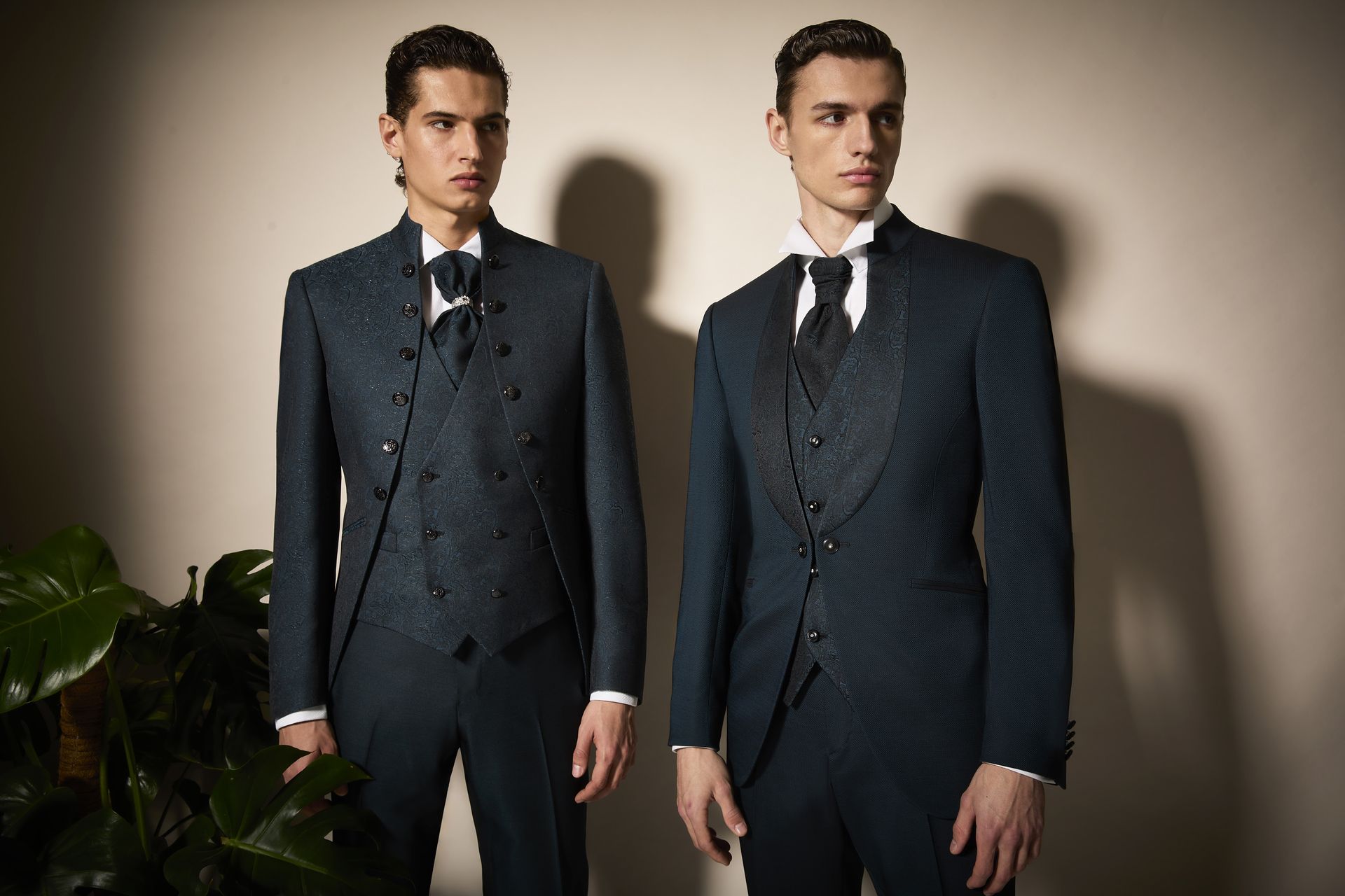 Due uomini in eleganti abiti color verde acqua sono in piedi vicino a una pianta frondosa, su uno sfondo color cuoio.