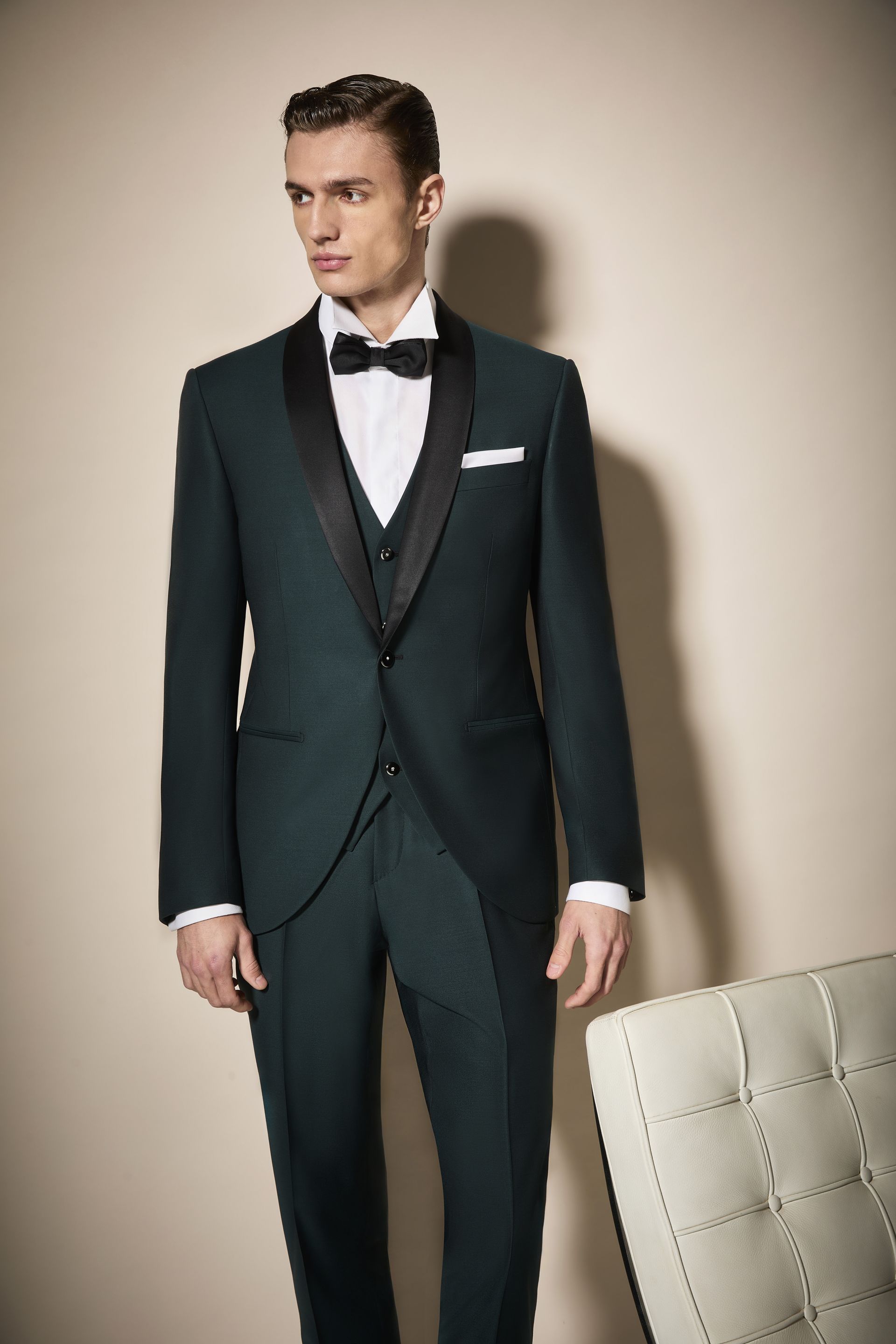 Uomo in smoking verde smeraldo con risvolti neri e papillon, in piedi in un interno vicino a una sedia.