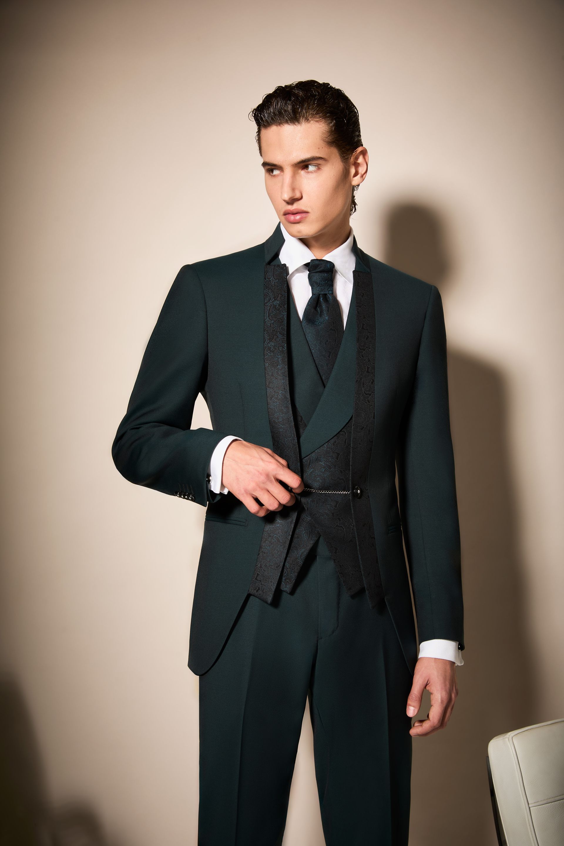 Uomo in abito formale verde scuro con cravatta nera e gilet, in posa in un interno.