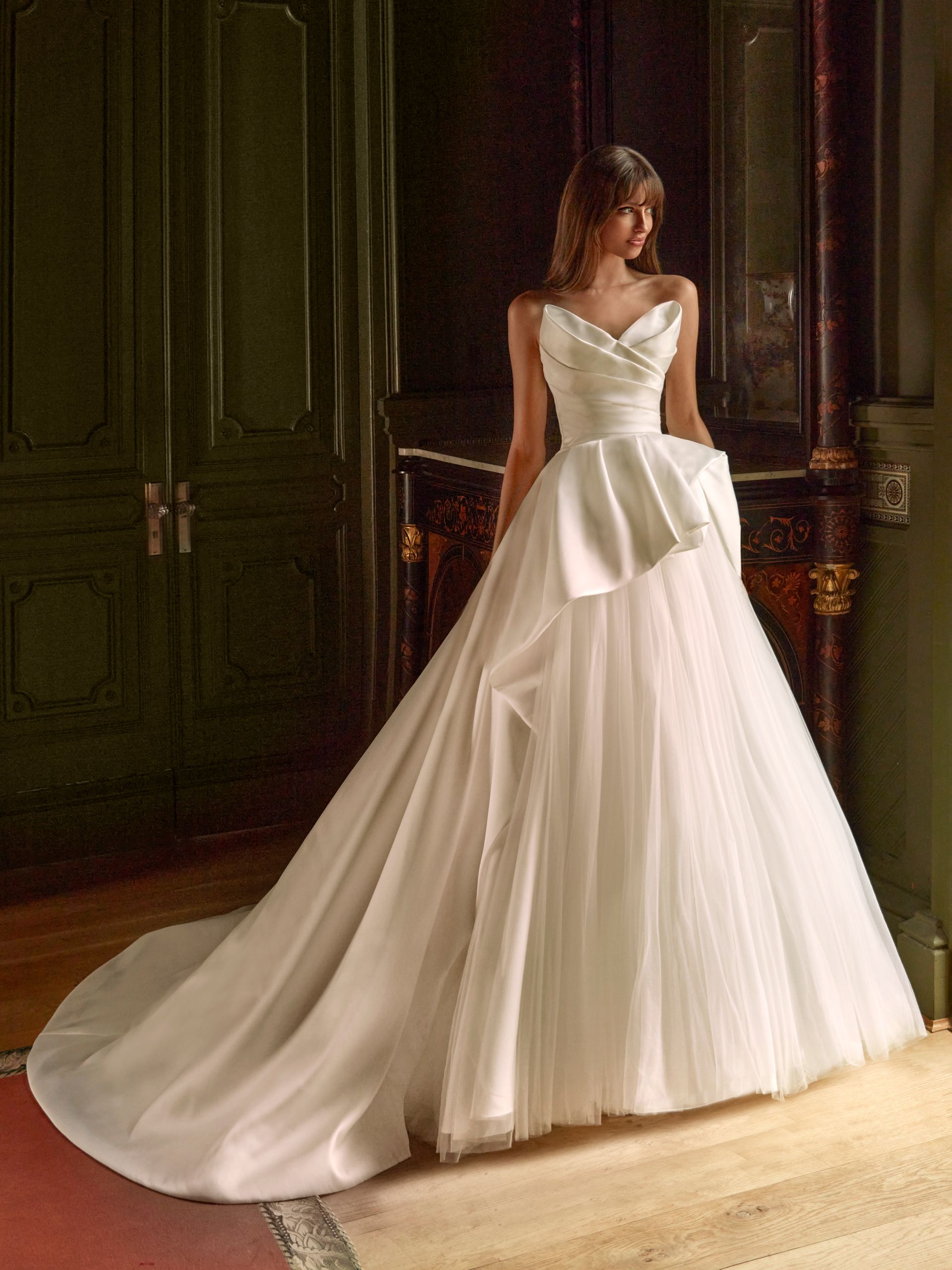 Donna in abito da sposa bianco con gonna a strati e strascico, in piedi in un interno.