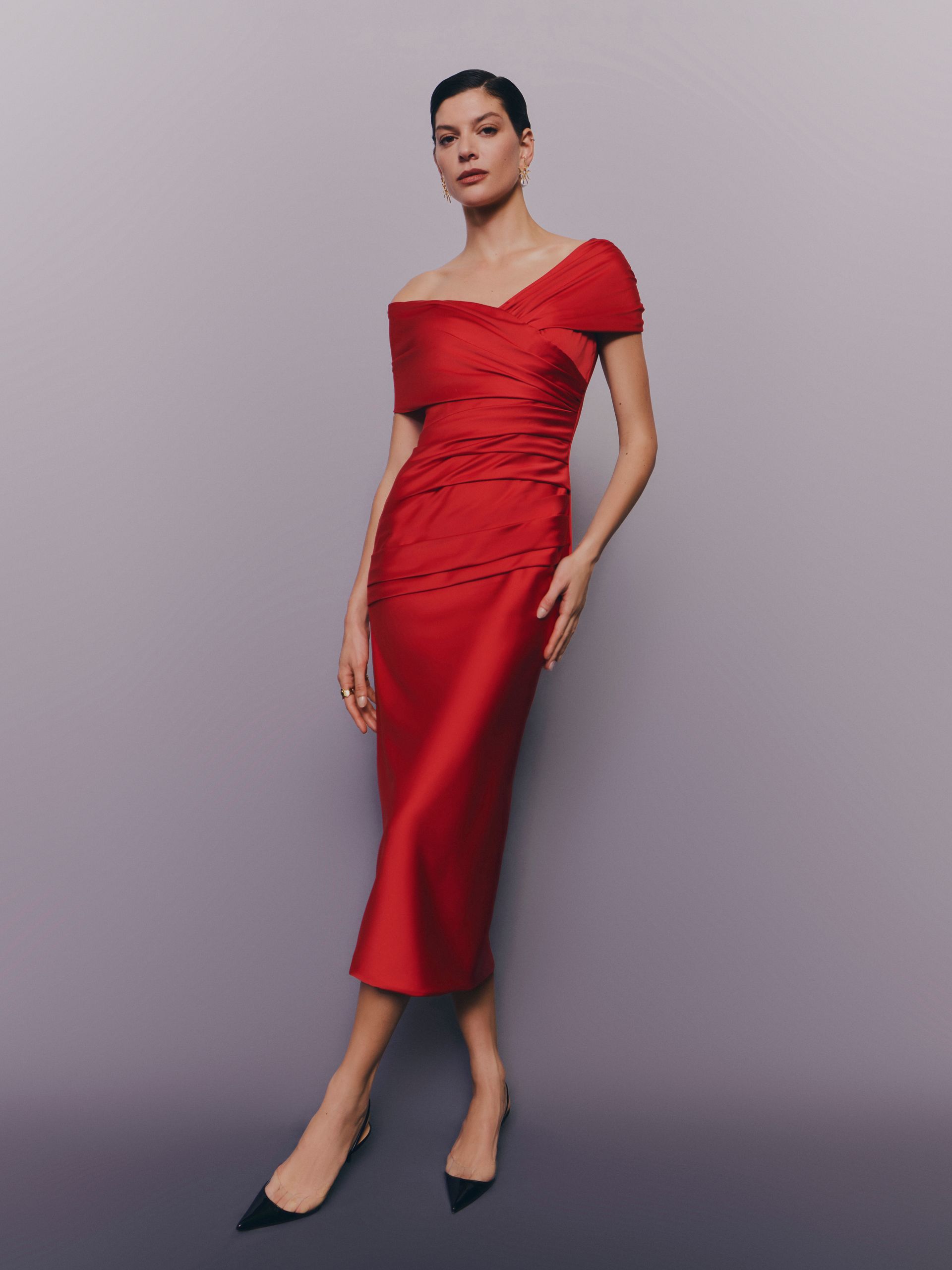 Una donna con un abito rosso a spalle scoperte posa su uno sfondo grigio e indossa tacchi neri.