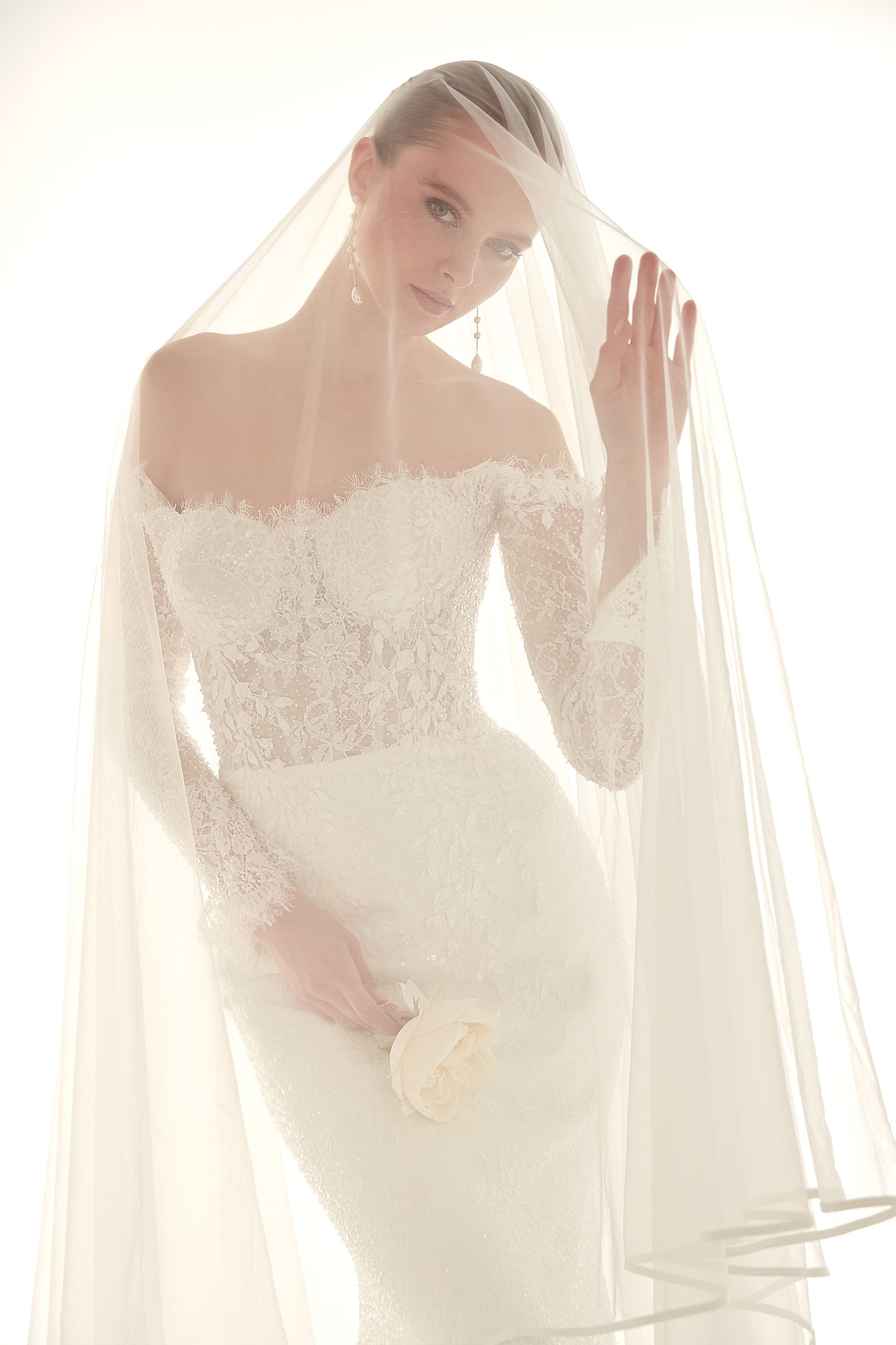 Donna con abito da sposa bianco in pizzo con spalle scoperte, velo e un fiore in mano.