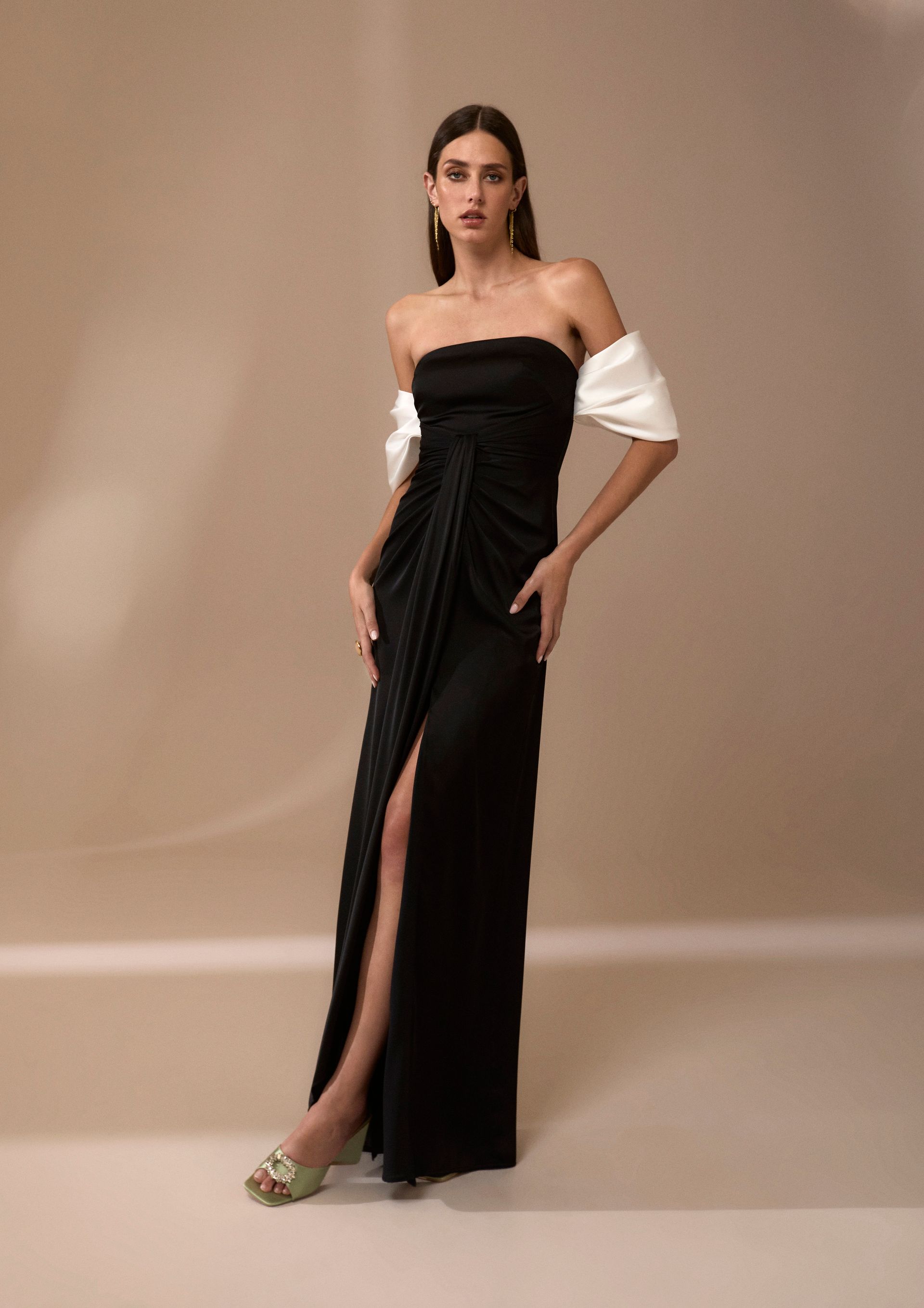 Una donna in abito nero senza spalline con spacco profondo, scialle bianco e orecchini d'oro posa su uno sfondo beige.