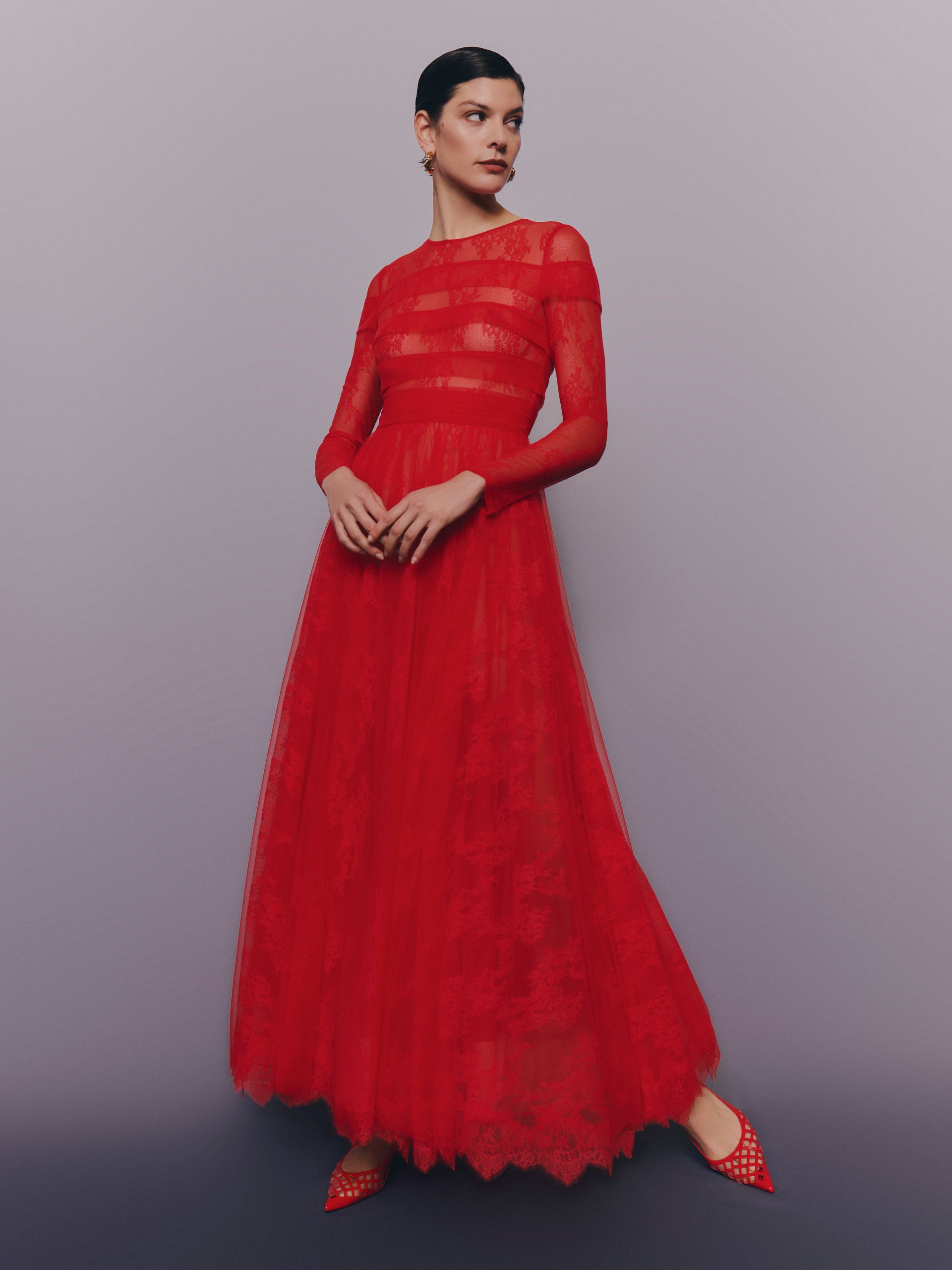Donna in un abito rosso lungo fino a terra, in piedi in uno studio. L'abito è a maniche lunghe, trasparente