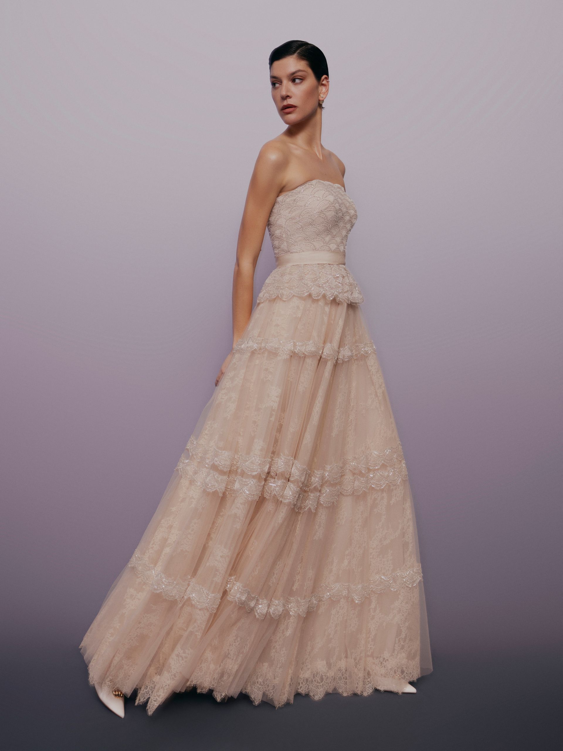 Donna con un abito a balze color cipria senza spalline, che guarda oltre la spalla, su uno sfondo sfumato lilla.