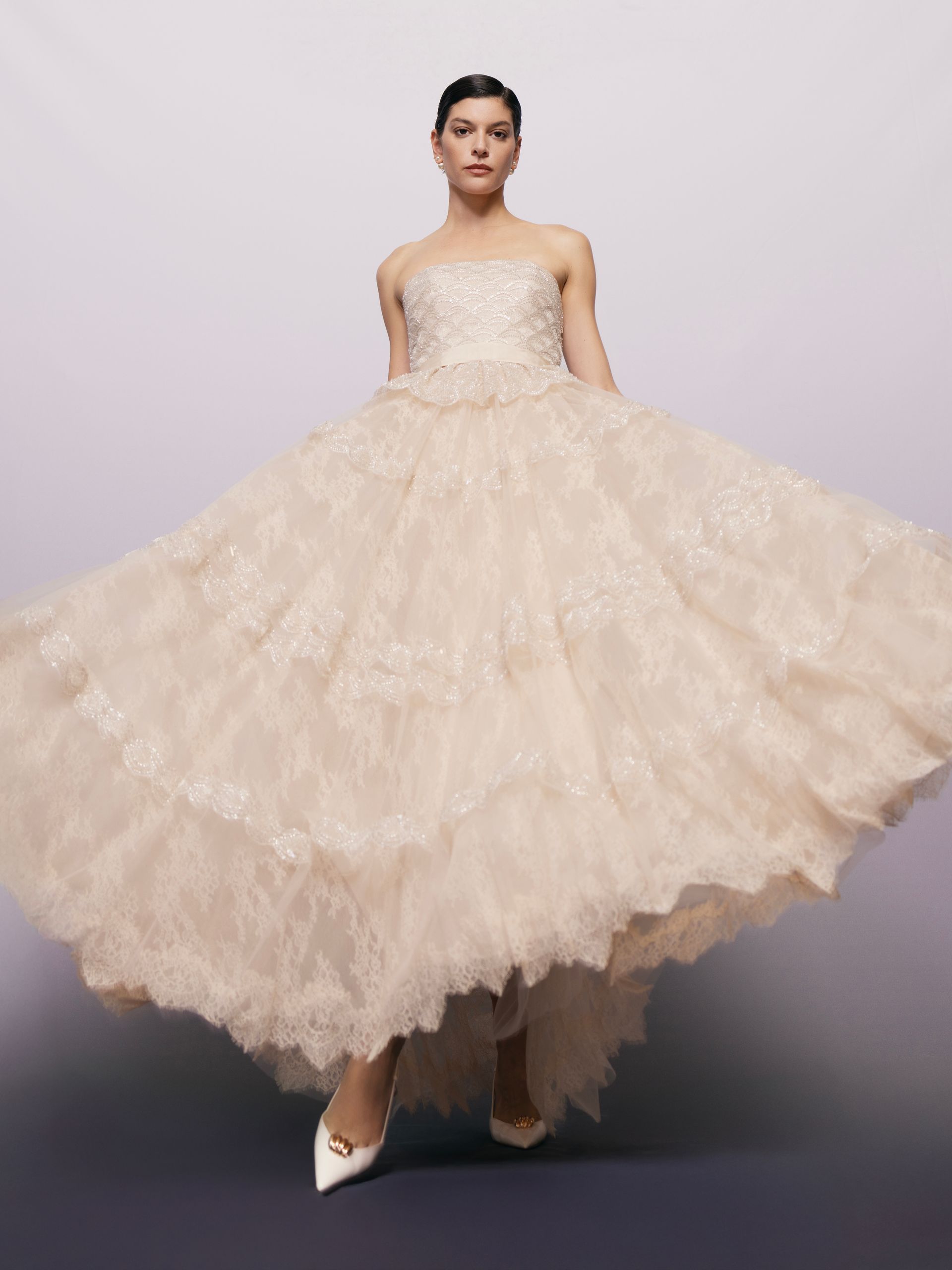 Donna in piedi, con indosso un abito da ballo rosa chiaro senza spalline, gonna voluminosa e scarpe bianche.