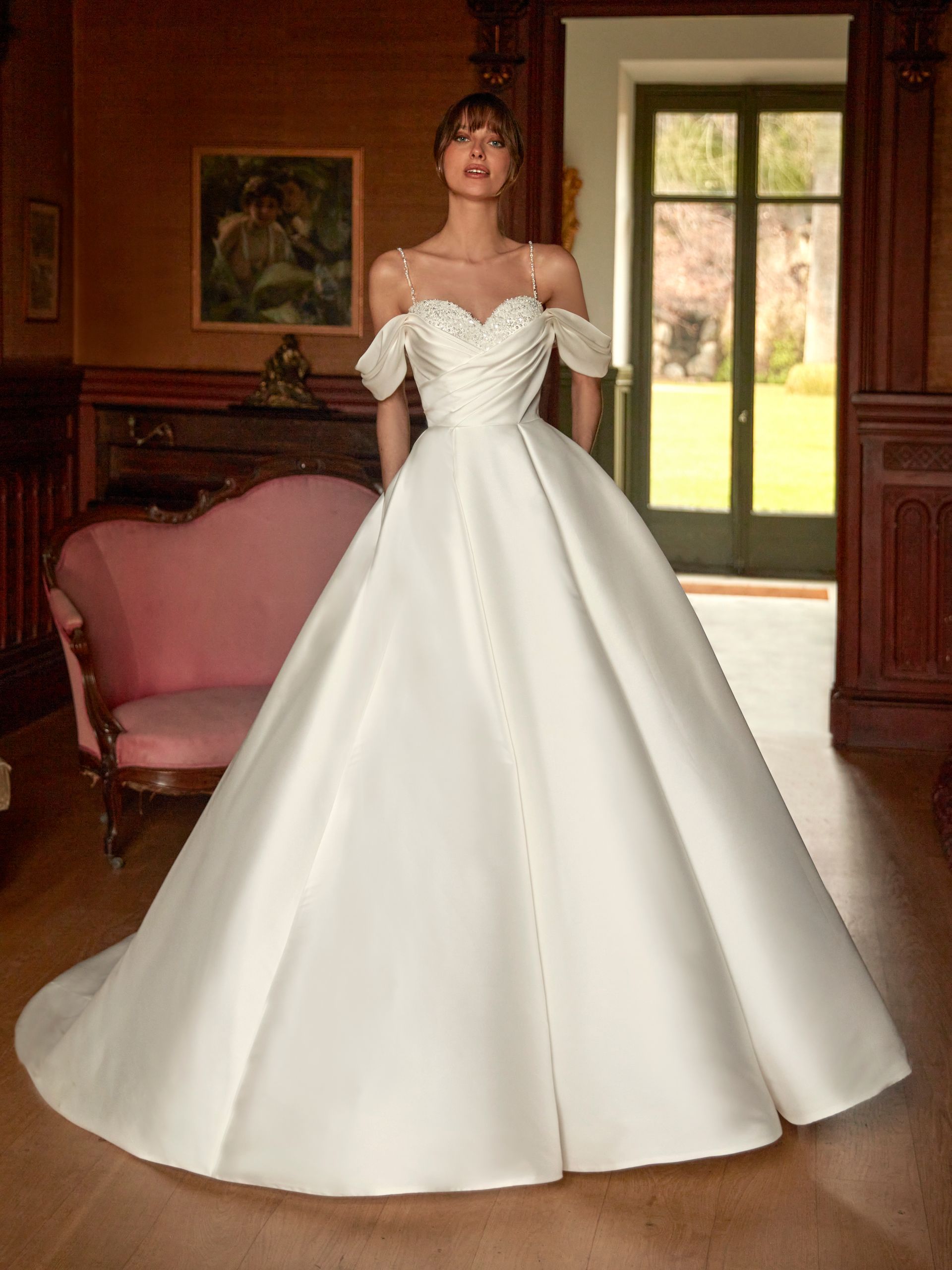 Donna in abito da sposa bianco con spalle scoperte, in piedi in un elegante ambiente interno.