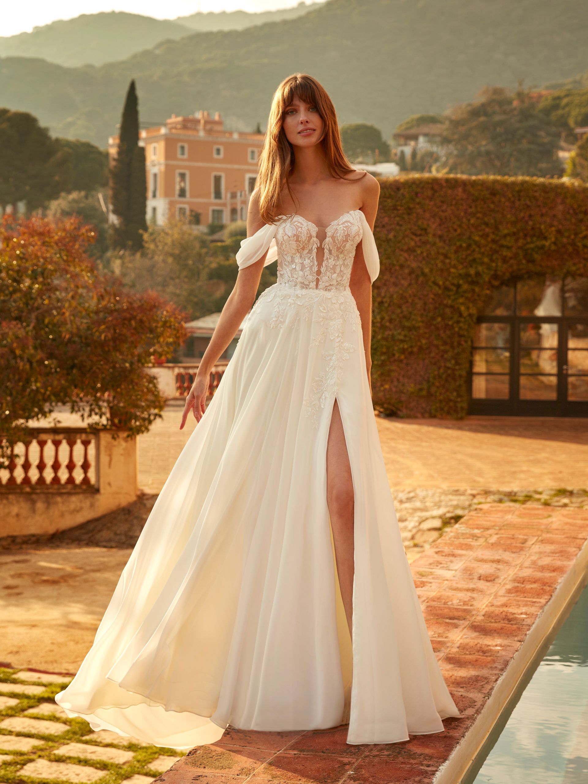 Donna in abito da sposa bianco con spalle scoperte e spacco fino alla coscia, in posa all'aperto.