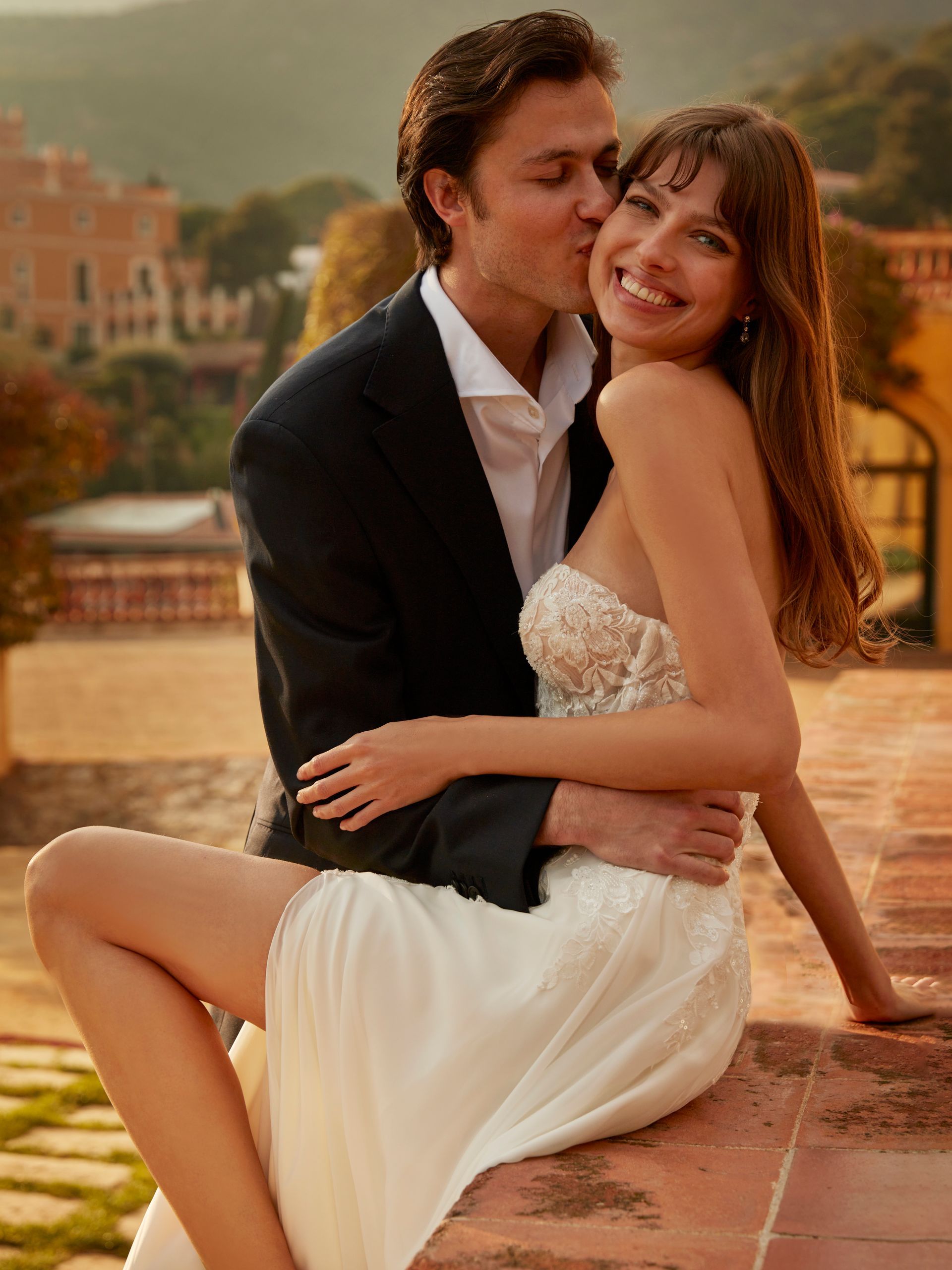 Un uomo bacia una donna sulla guancia; siedono insieme, sorridenti, su una terrazza in pietra. 