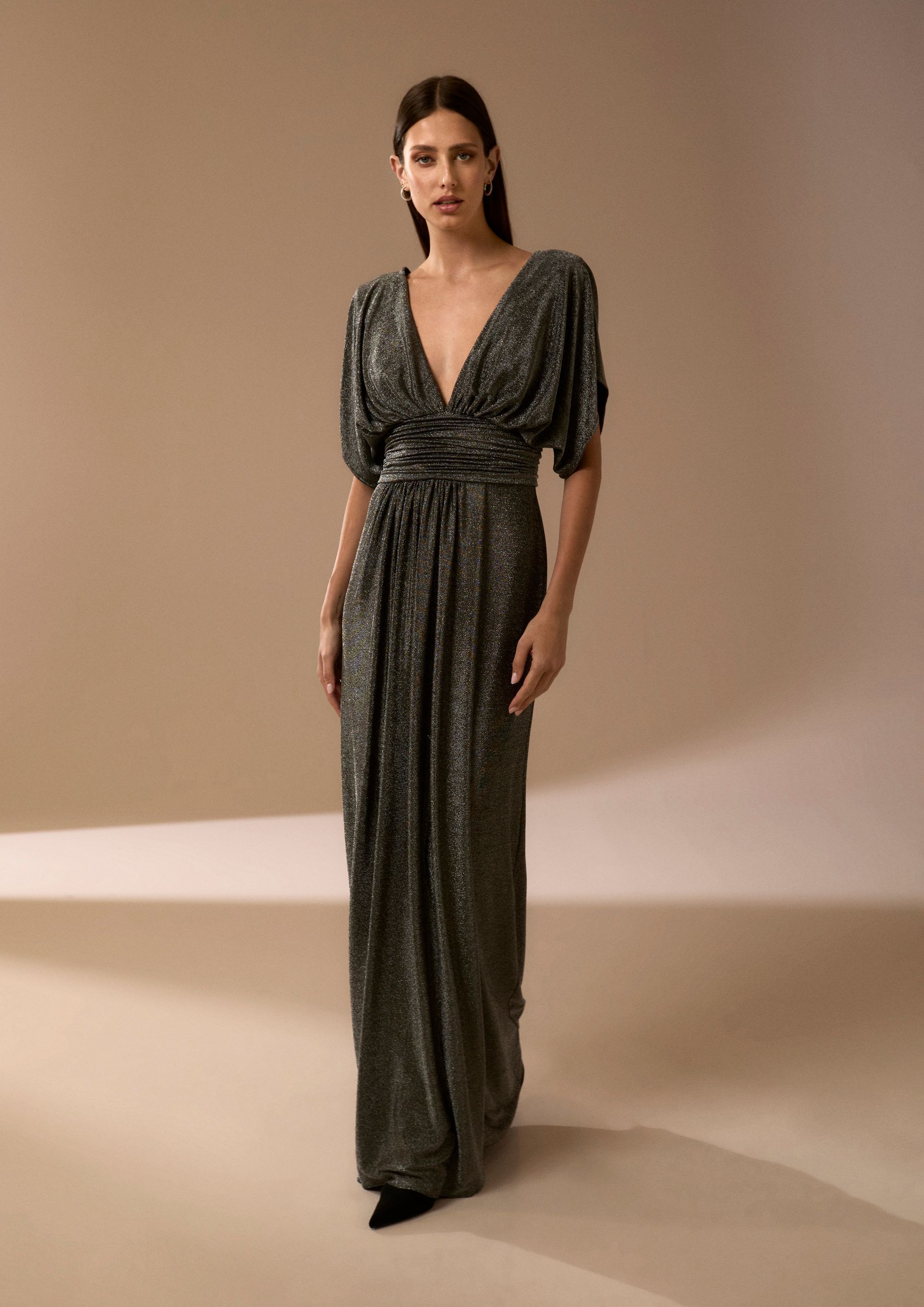 Donna in piedi in un interno, con indosso un abito metallico lungo fino ai piedi, con profonda scollatura a V e maniche corte.