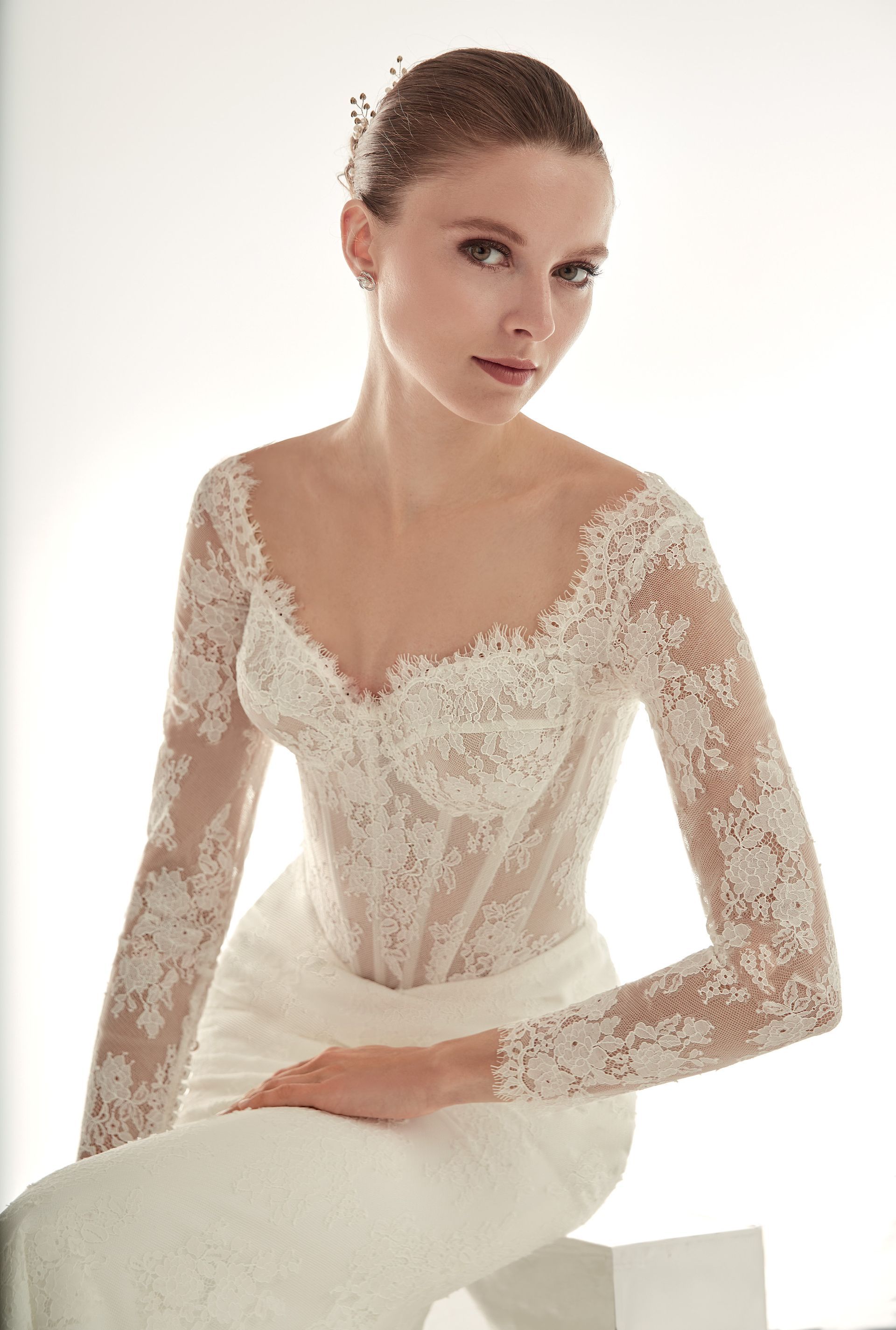 Donna seduta con abito da sposa in pizzo bianco, maniche lunghe, scollatura profonda, che guarda la telecamera.