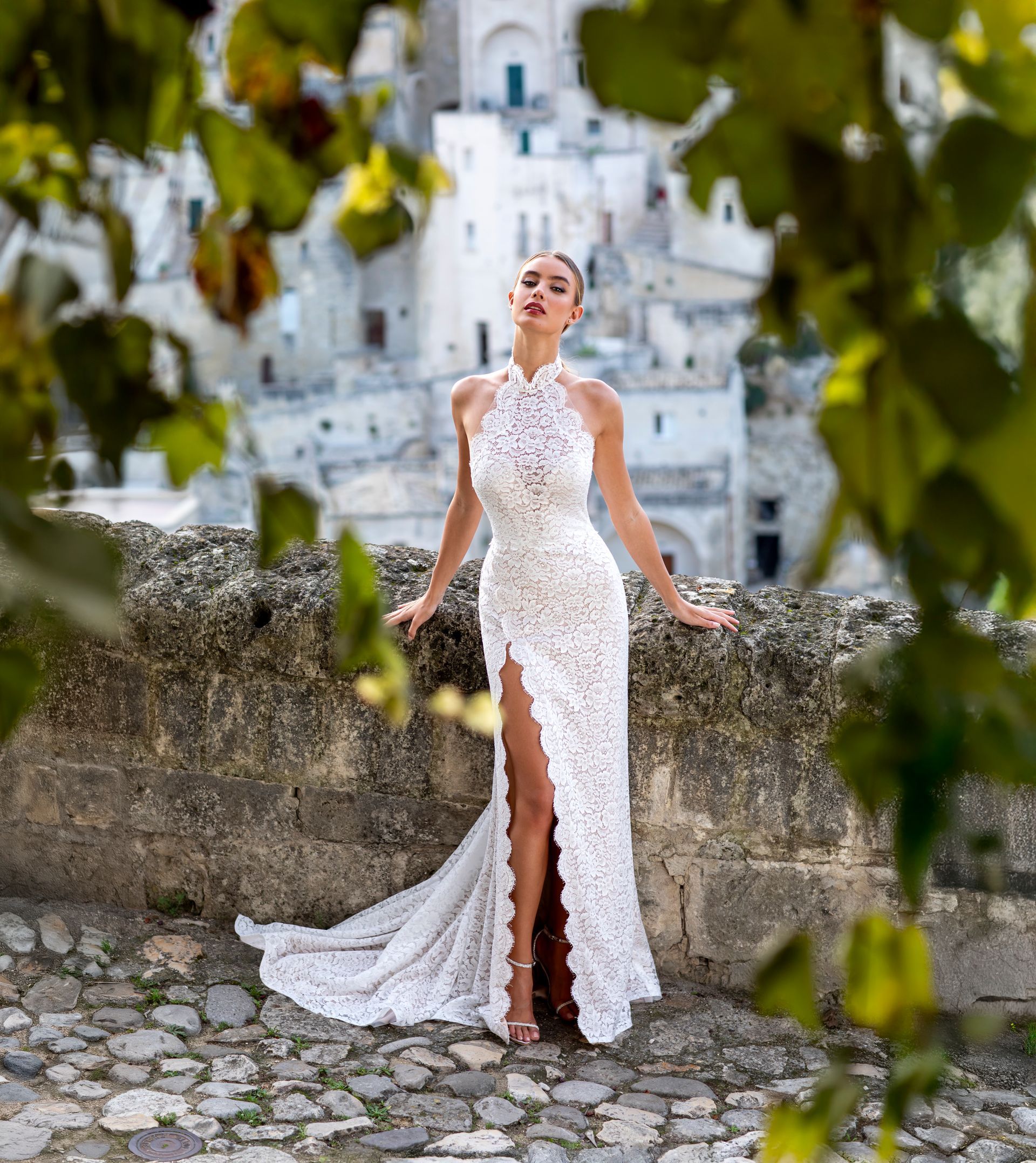 Una donna in abito di pizzo bianco con spacco profondo posa all'aperto; sullo sfondo una città storica.