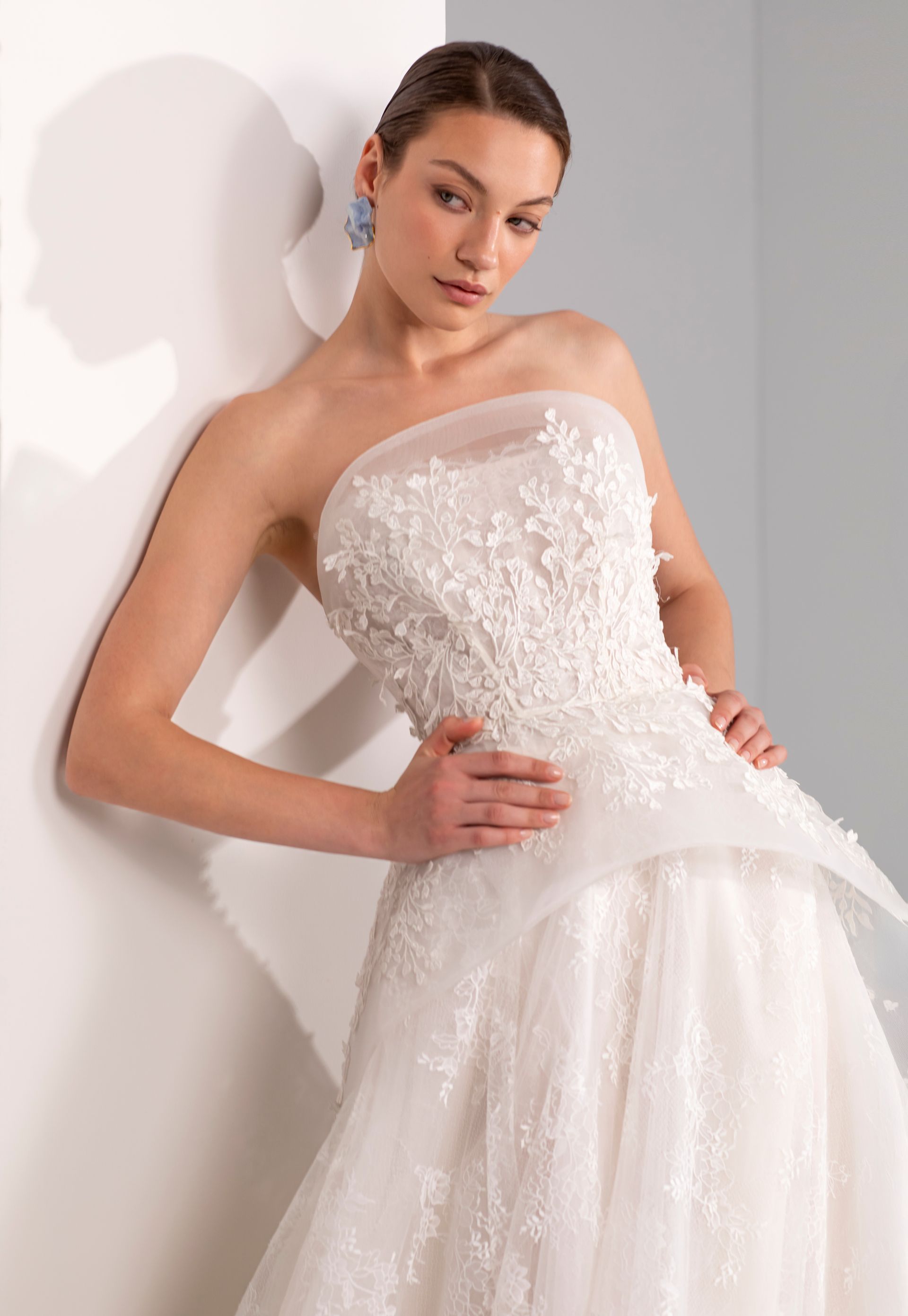Una donna con un abito da sposa bianco senza spalline è appoggiata a un muro, con una mano sul fianco.