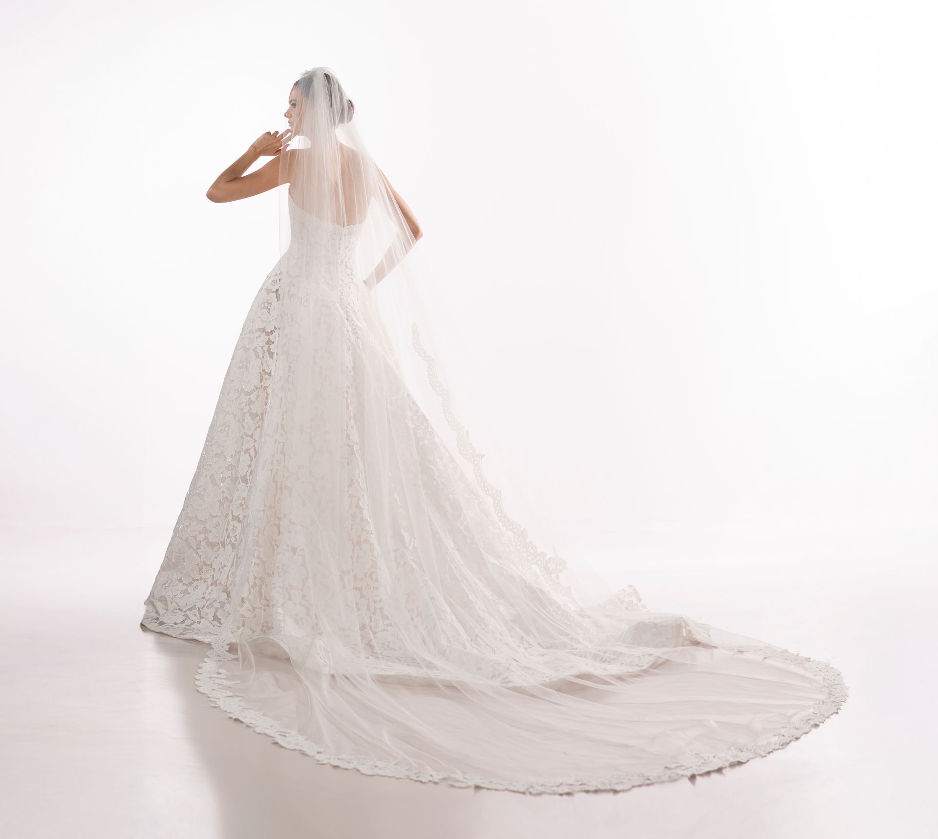 Sposa in abito bianco e lungo velo, di spalle, su sfondo bianco.