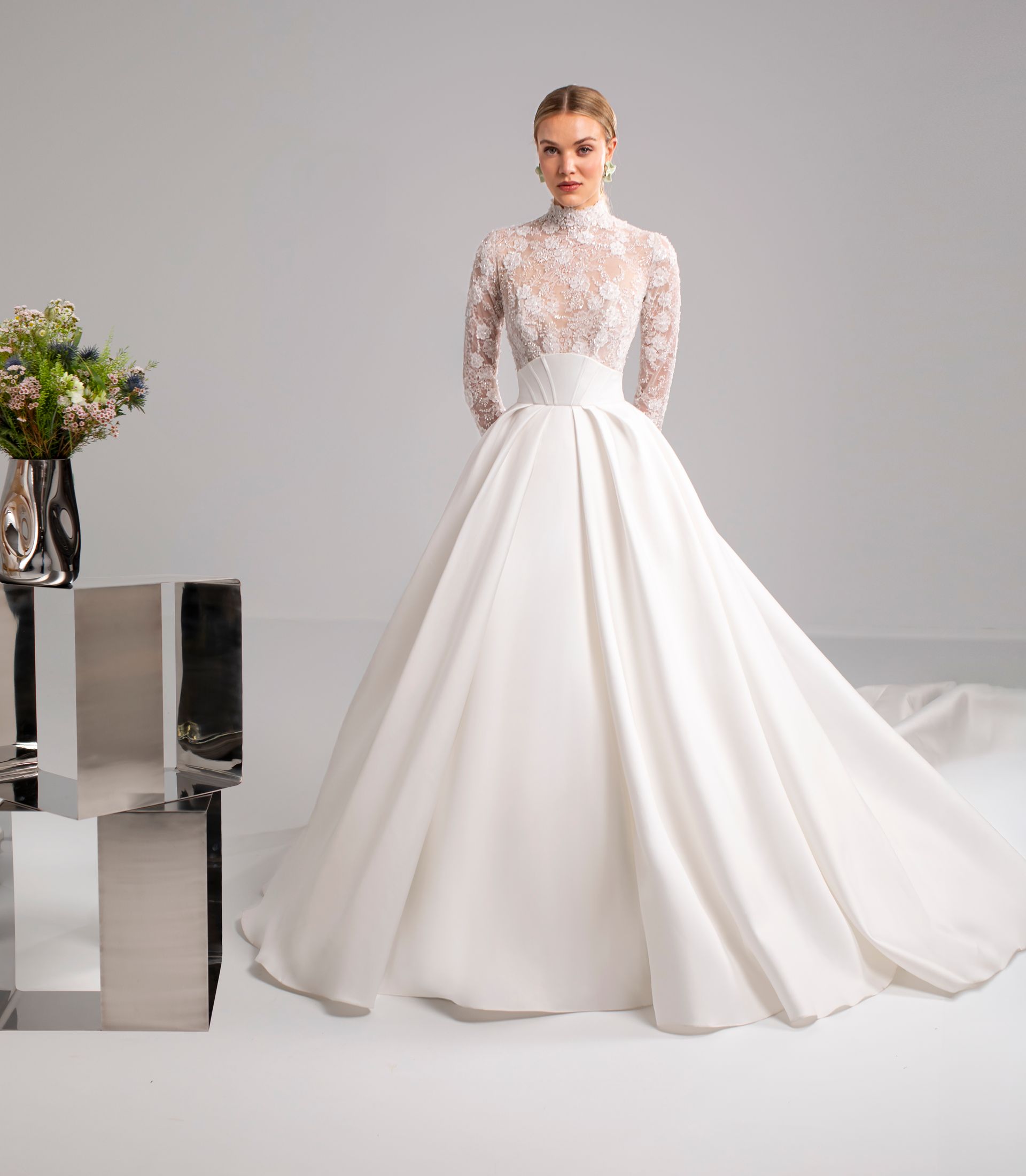 Donna in abito da sposa bianco, maniche lunghe, collo alto, gonna ampia, in piedi in uno studio.