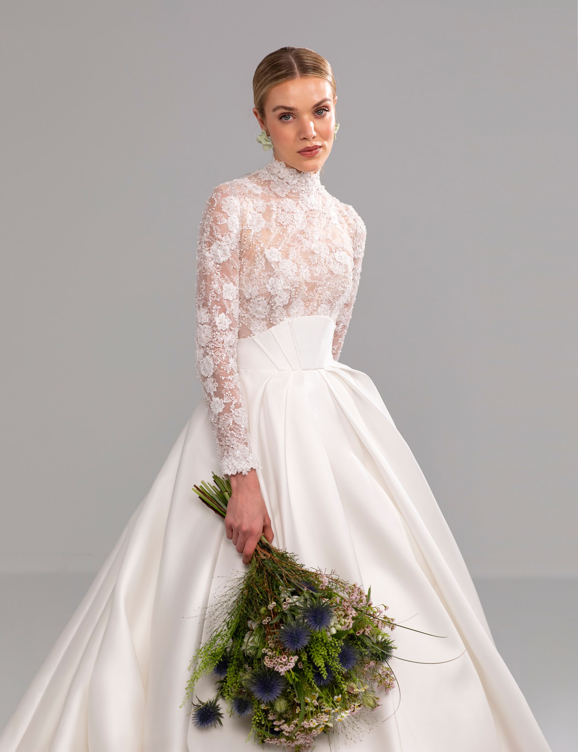 Donna in abito da sposa bianco con lunghe maniche in pizzo, che tiene in mano un bouquet, su uno sfondo grigio.