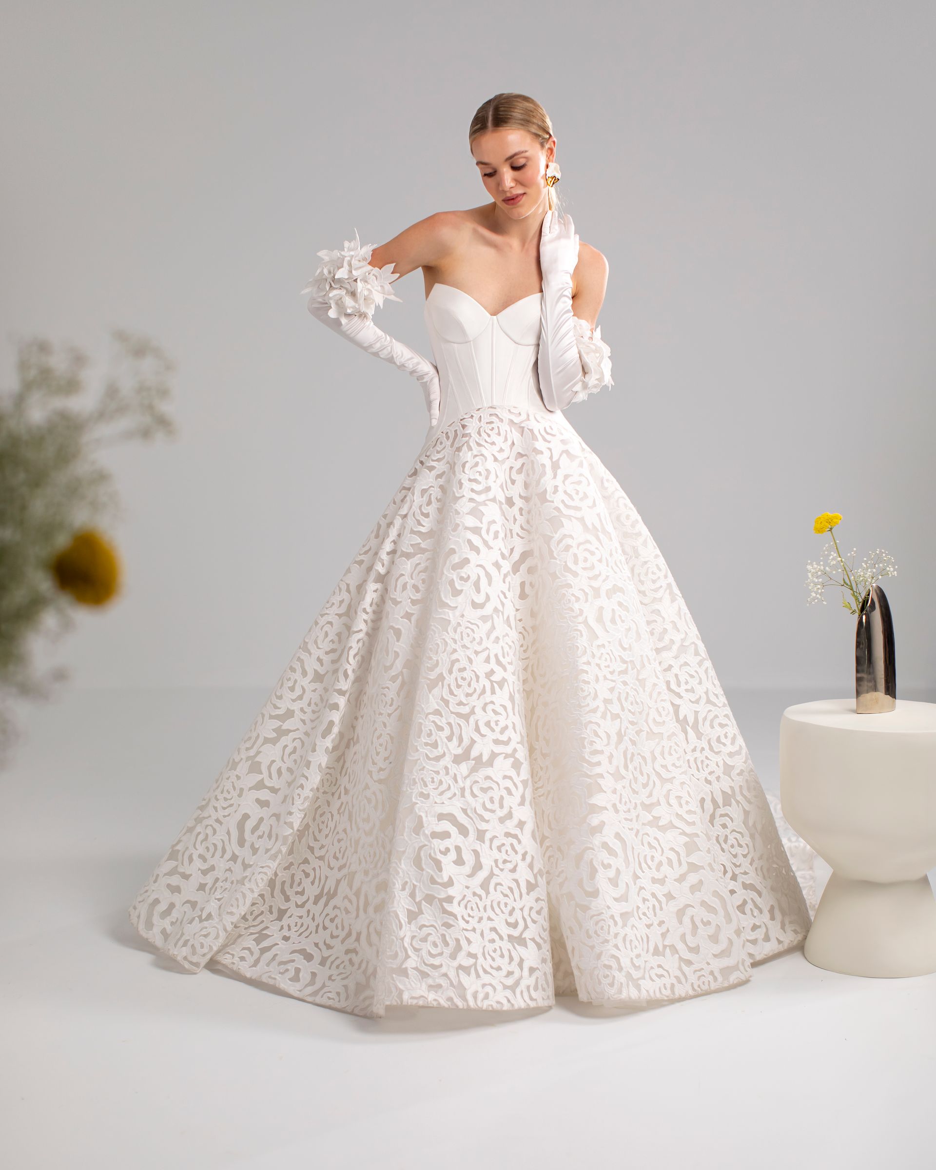 Sposa sorridente in abito di pizzo senza spalline con guanti lunghi. Sfondo bianco, piccoli dettagli floreali.