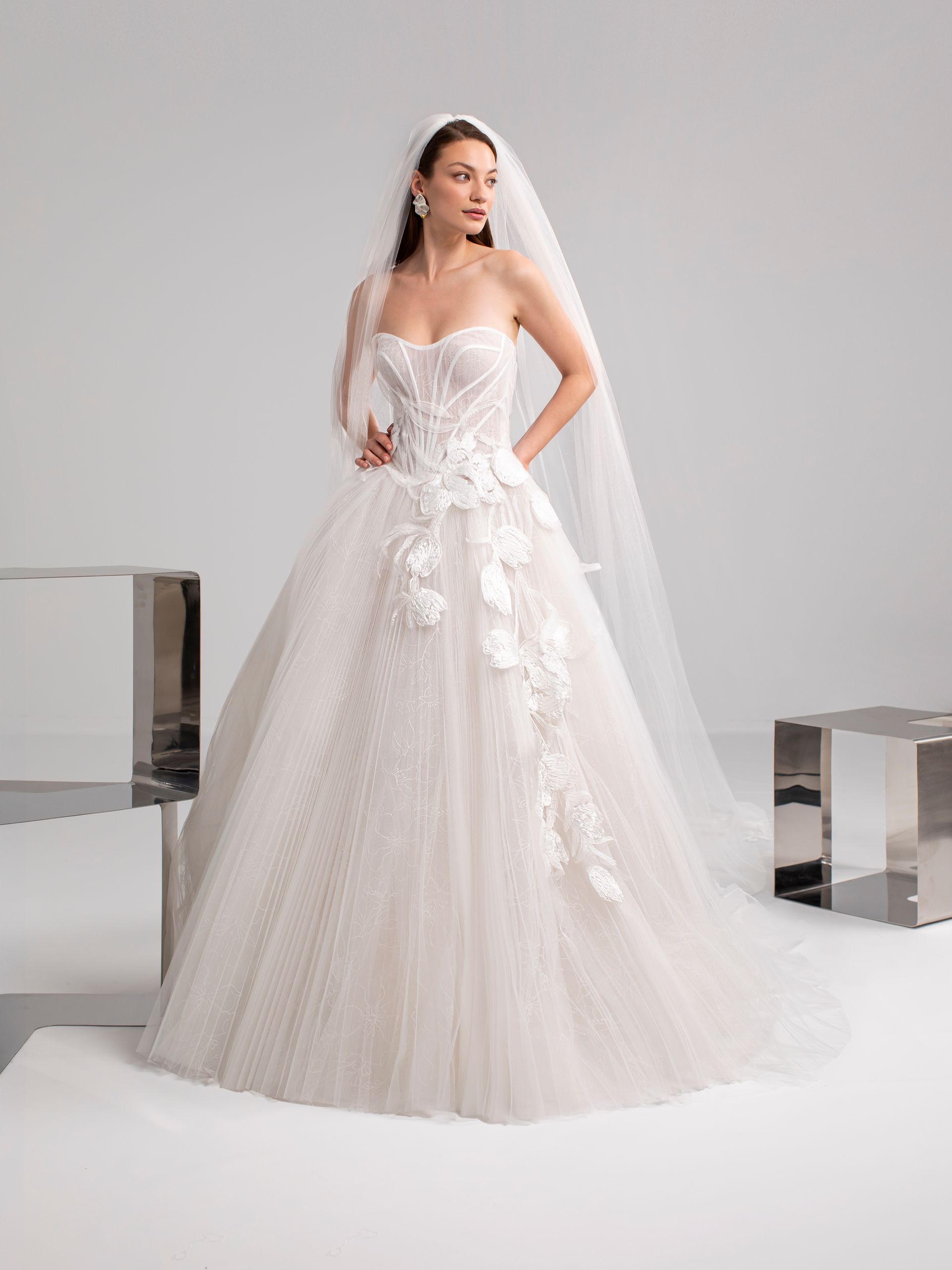 Donna in abito da sposa bianco senza spalline, con decorazioni floreali e velo, in posa su uno sfondo bianco.