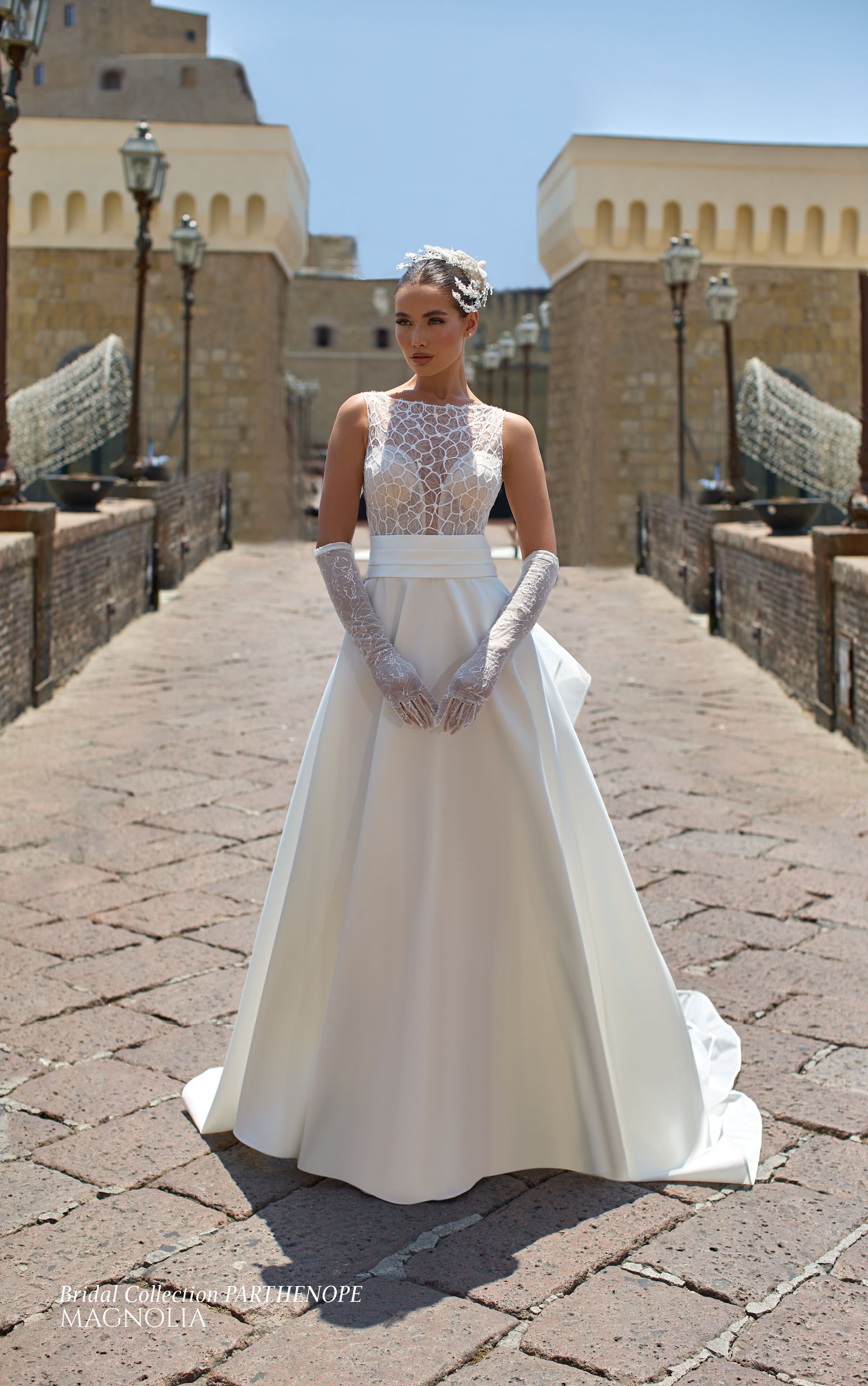 Una donna in abito da sposa bianco e guanti è in piedi su un ponte di pietra, sorridente, con un castello sullo sfondo.