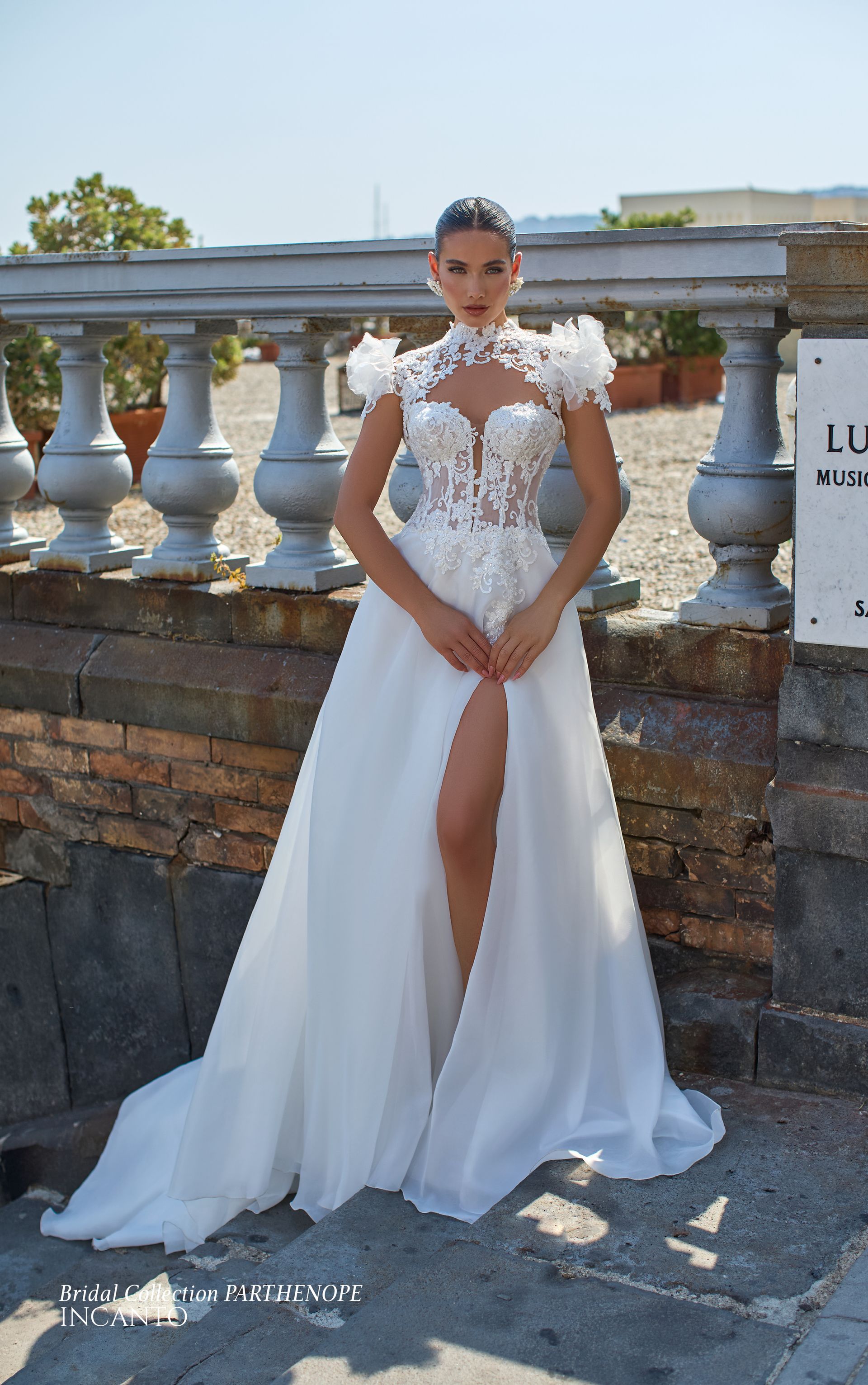 Una donna in abito da sposa bianco con spacco profondo e corpetto floreale è in piedi all'aperto vicino a un muro di pietra.