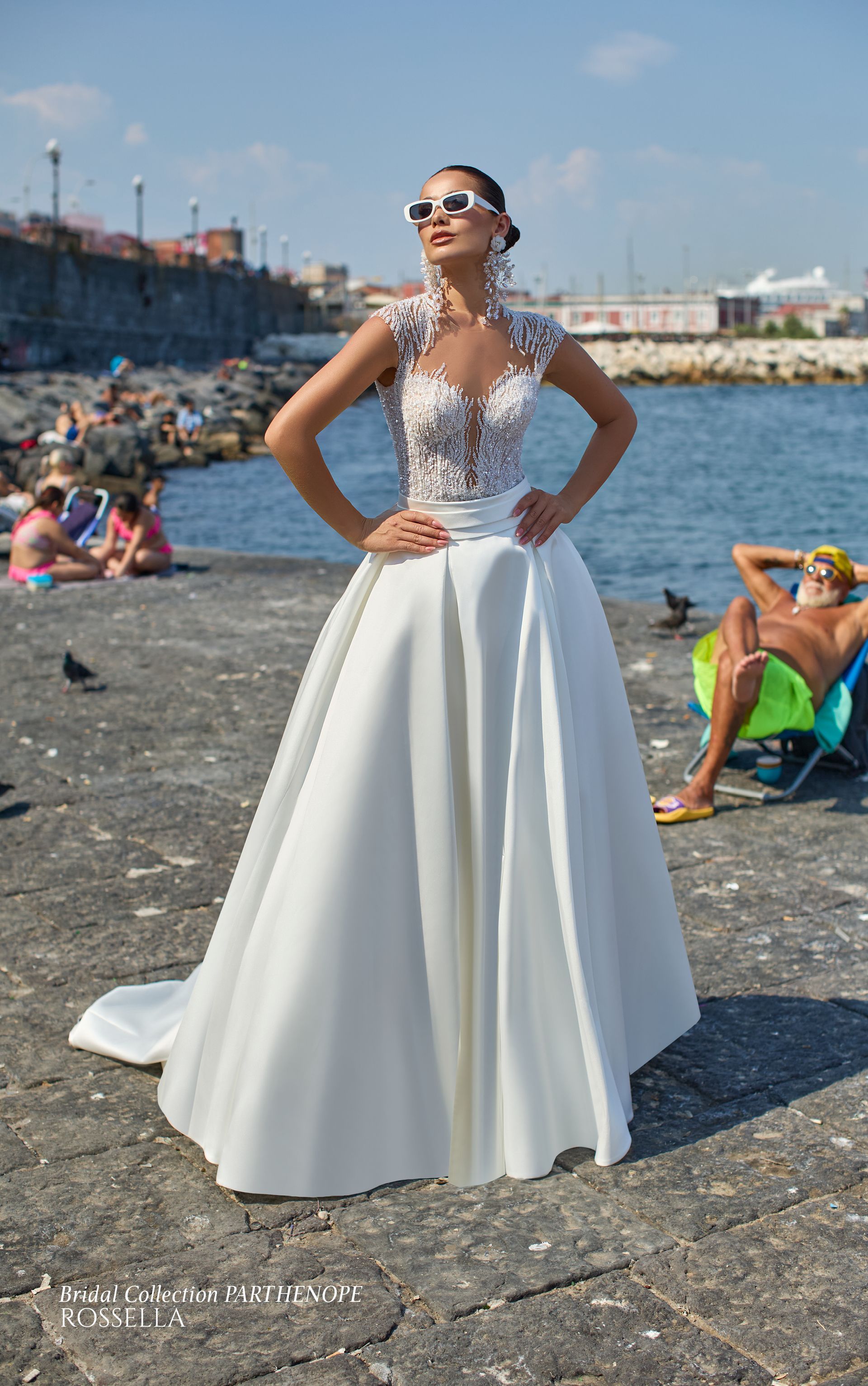 Donna in abito da sposa bianco con corpetto floreale, mani sui fianchi, in piedi in riva al mare con occhiali da sole.