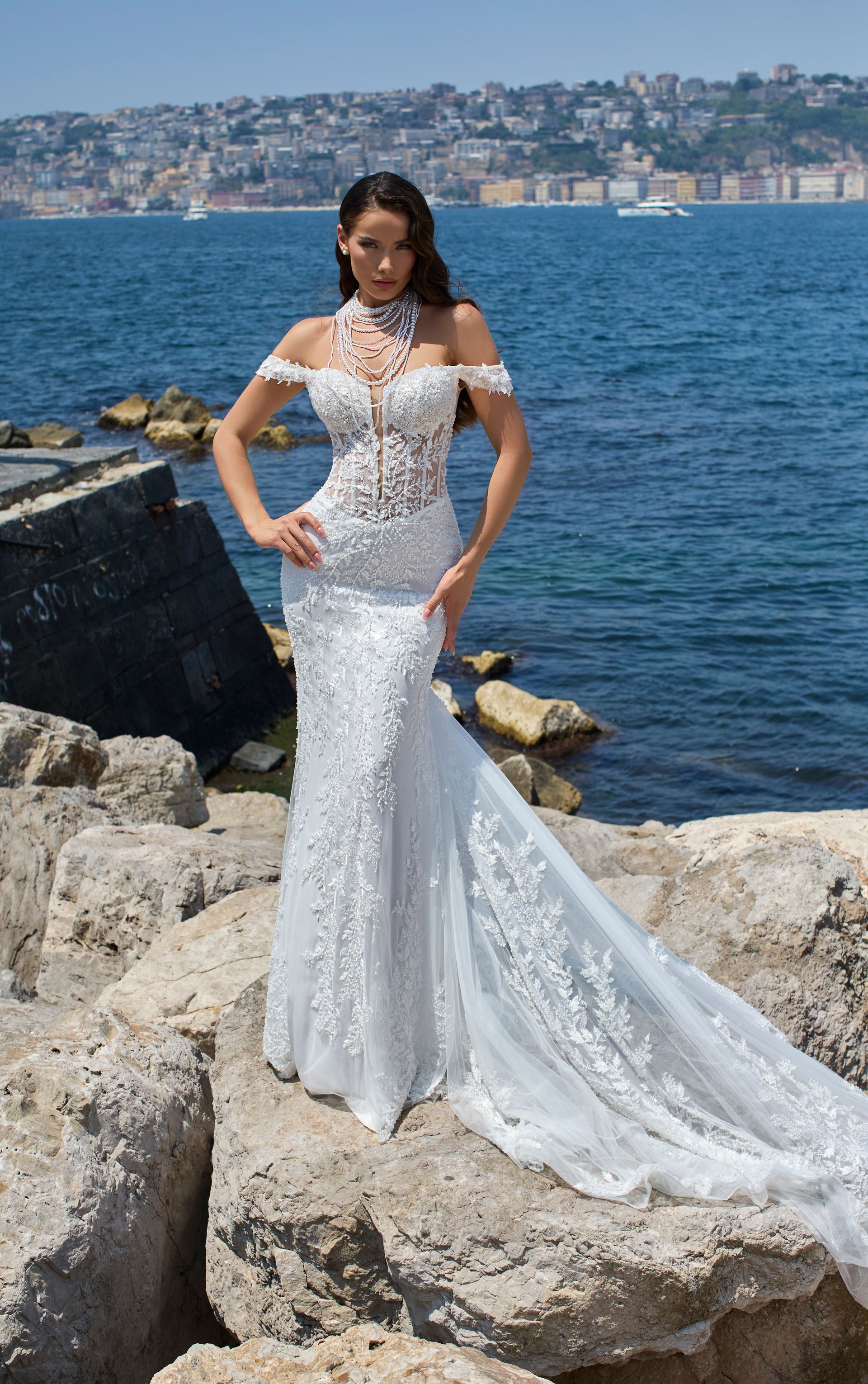 Donna in abito da sposa bianco con le spalle scoperte, sulle rocce in riva al mare, con le mani sui fianchi, che guarda.