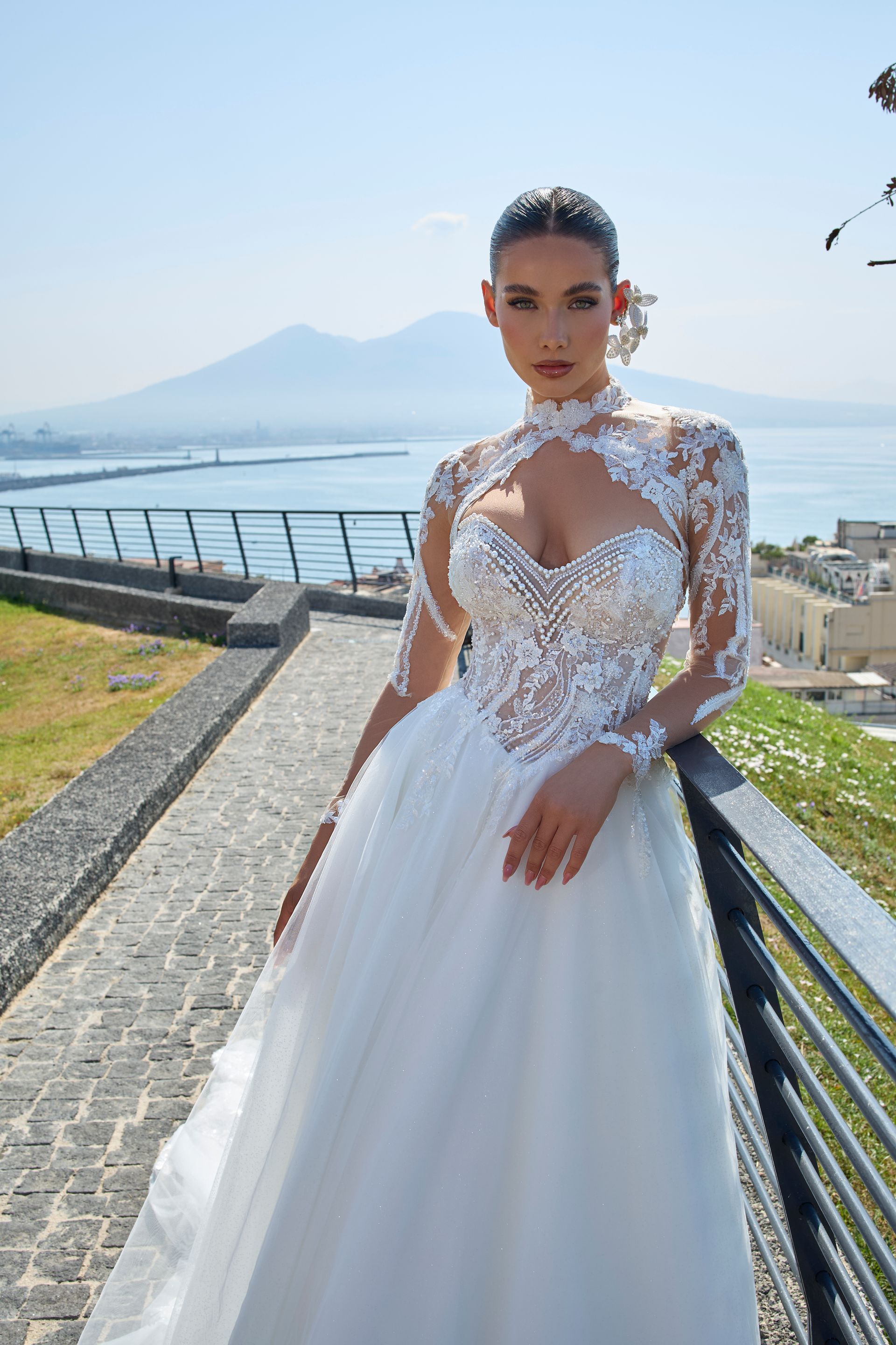 Donna in abito da sposa bianco con maniche in pizzo, in posa all'aperto con montagne e mare sullo sfondo.