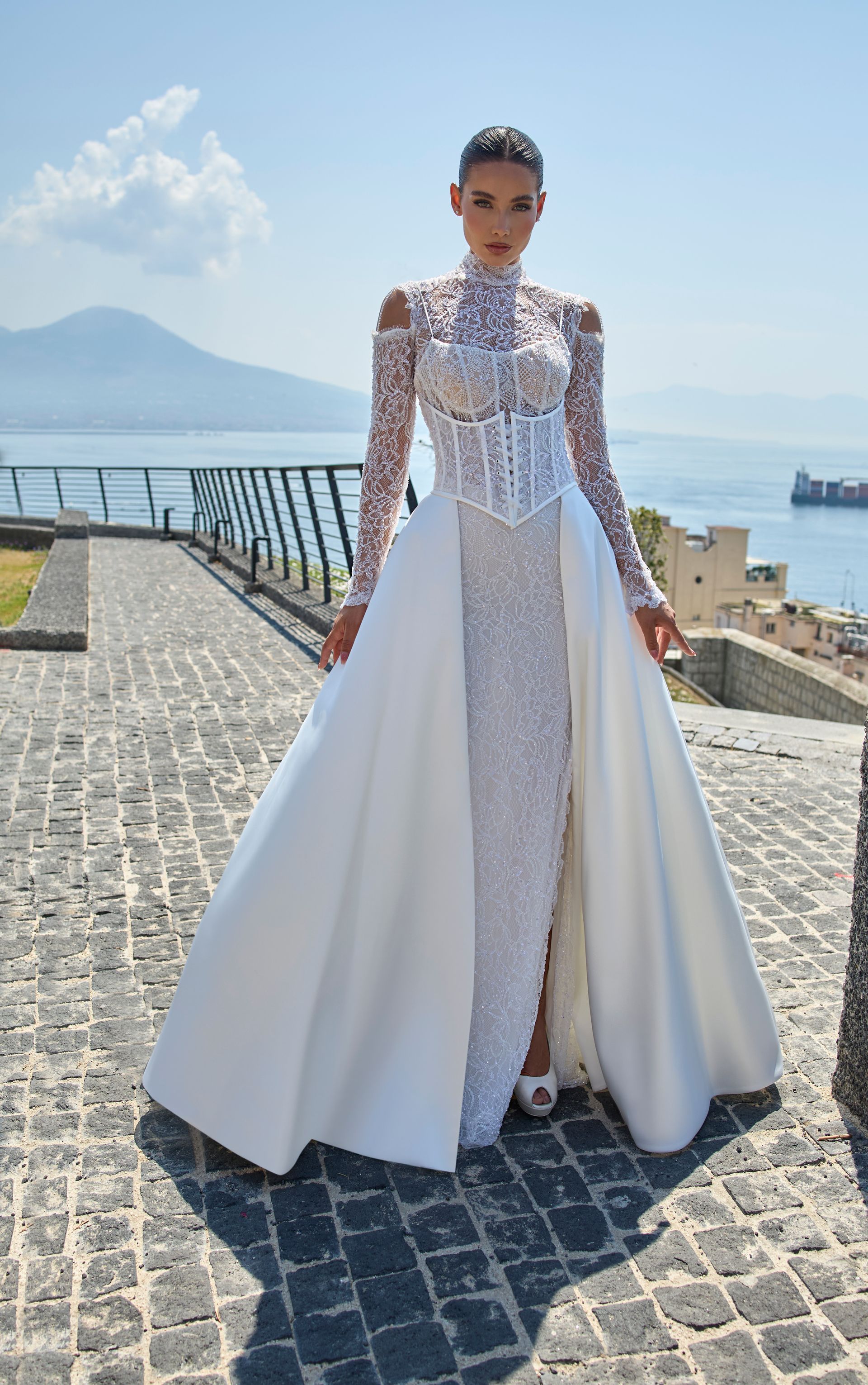 Donna in abito formale bianco con lungo strascico, su una passerella in pietra con vista sul mare