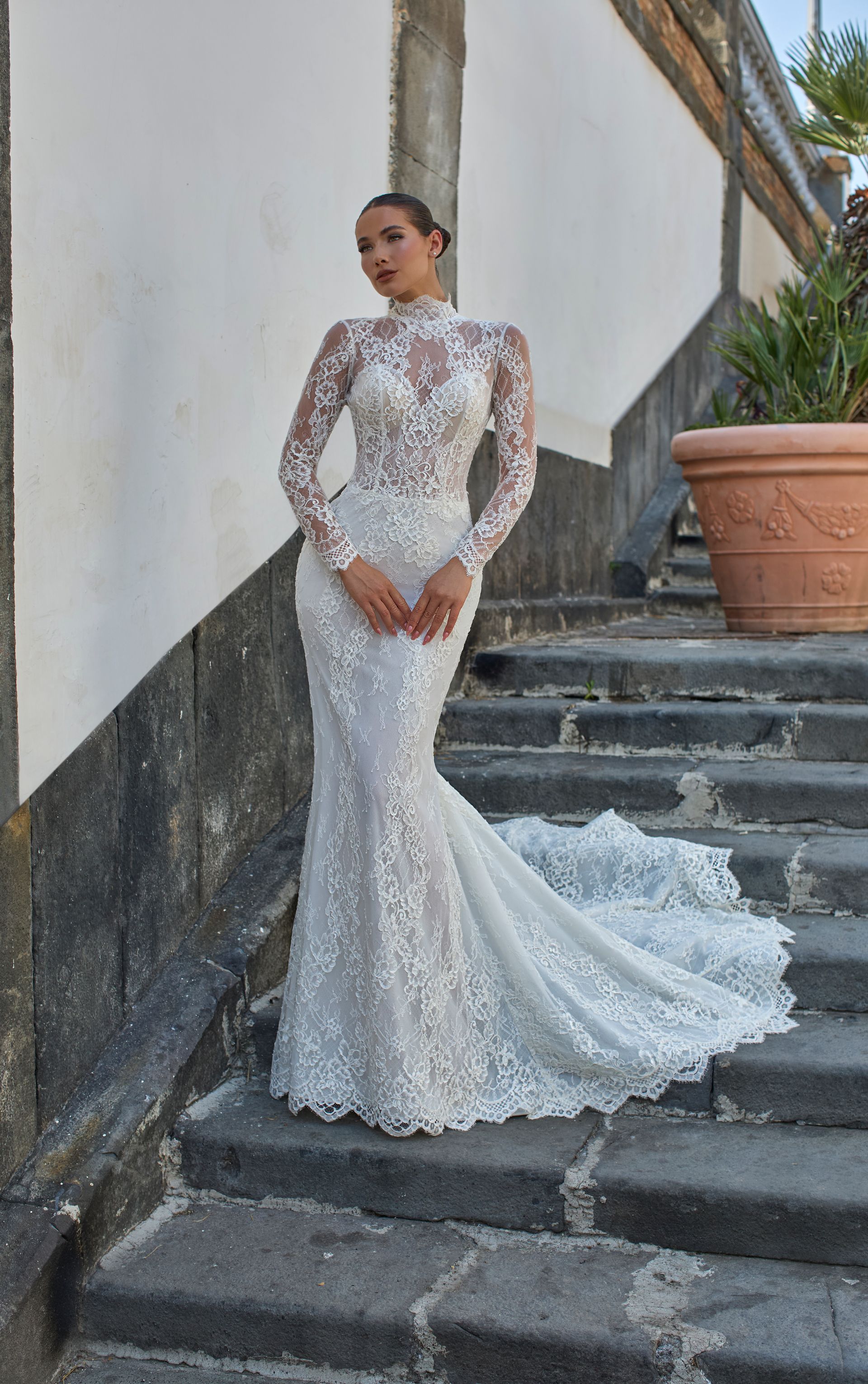 Donna in abito da sposa in pizzo bianco con maniche lunghe e strascico, su gradini di pietra.