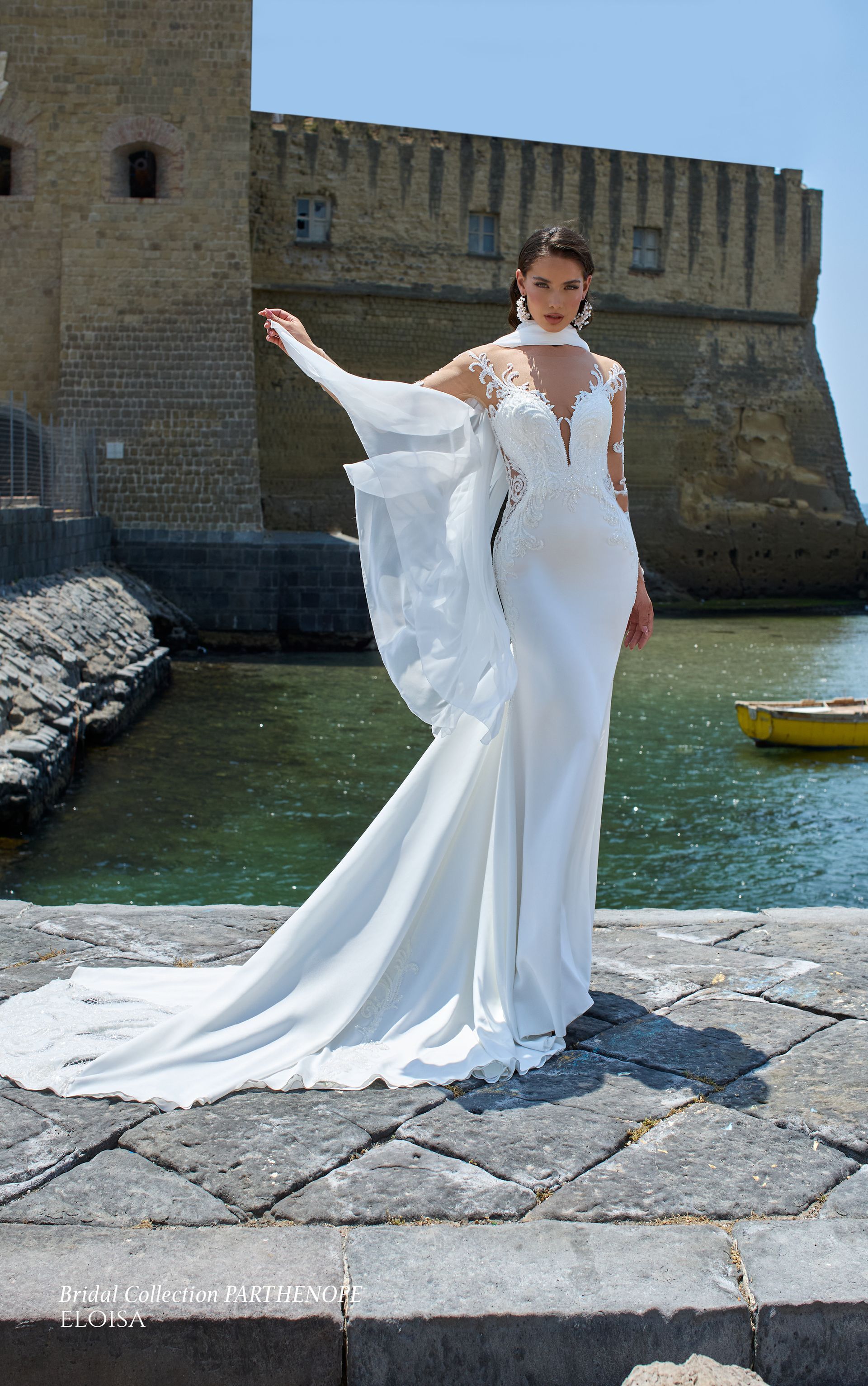 Donna in abito da sposa bianco con strascico e mantello trasparente, in piedi vicino a un muro di pietra e all'acqua.