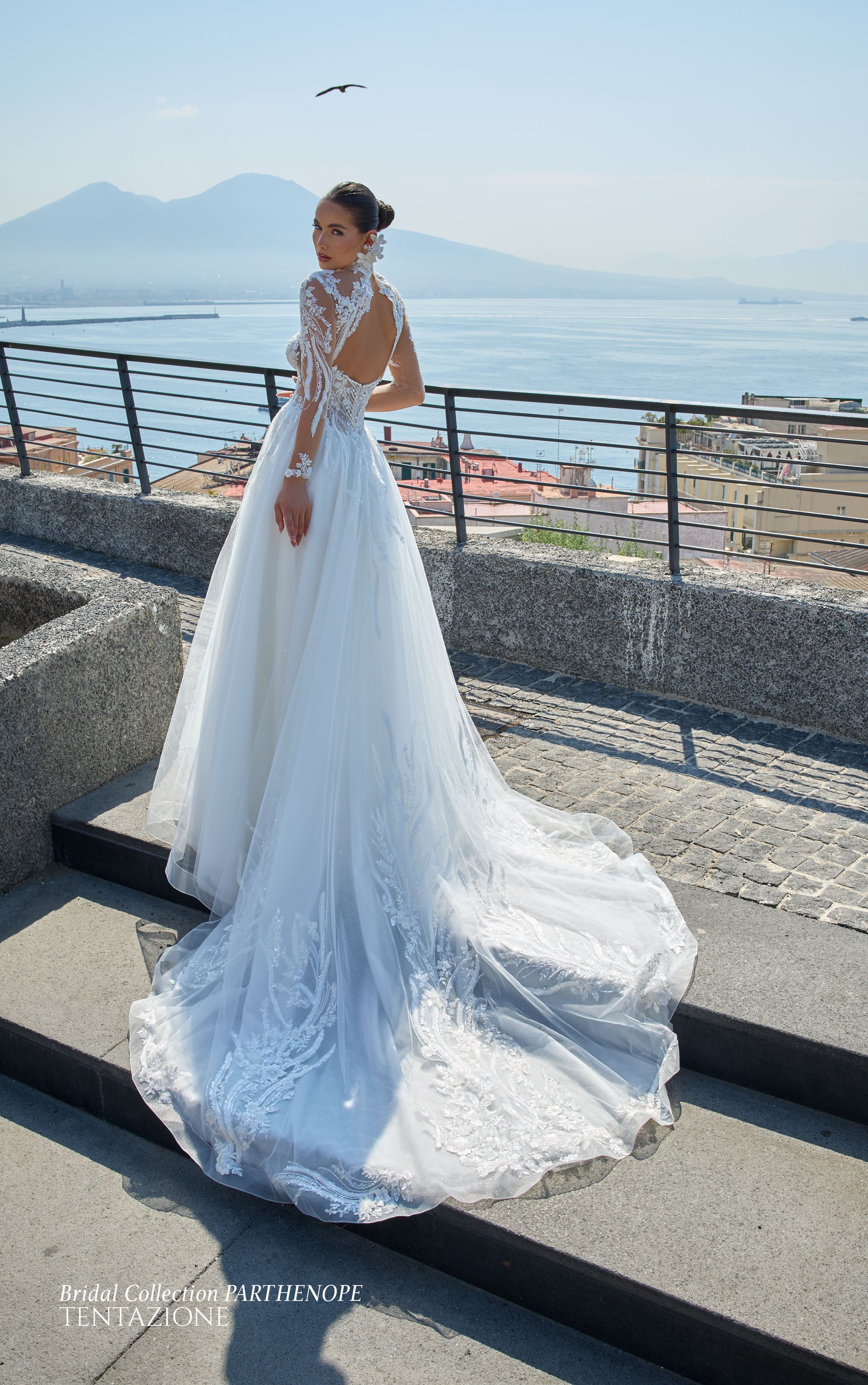 Donna in abito da sposa bianco con lungo strascico, in piedi sui gradini con vista sul lungomare.