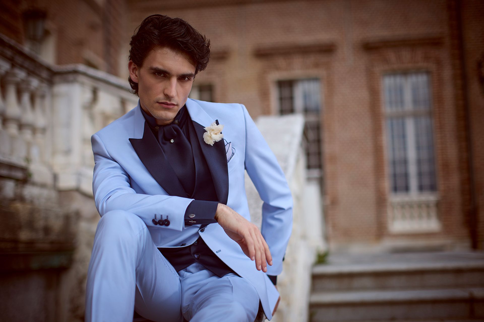 Uomo in abito azzurro, seduto sulle scale, in posa con un'espressione seria, ambiente formale.