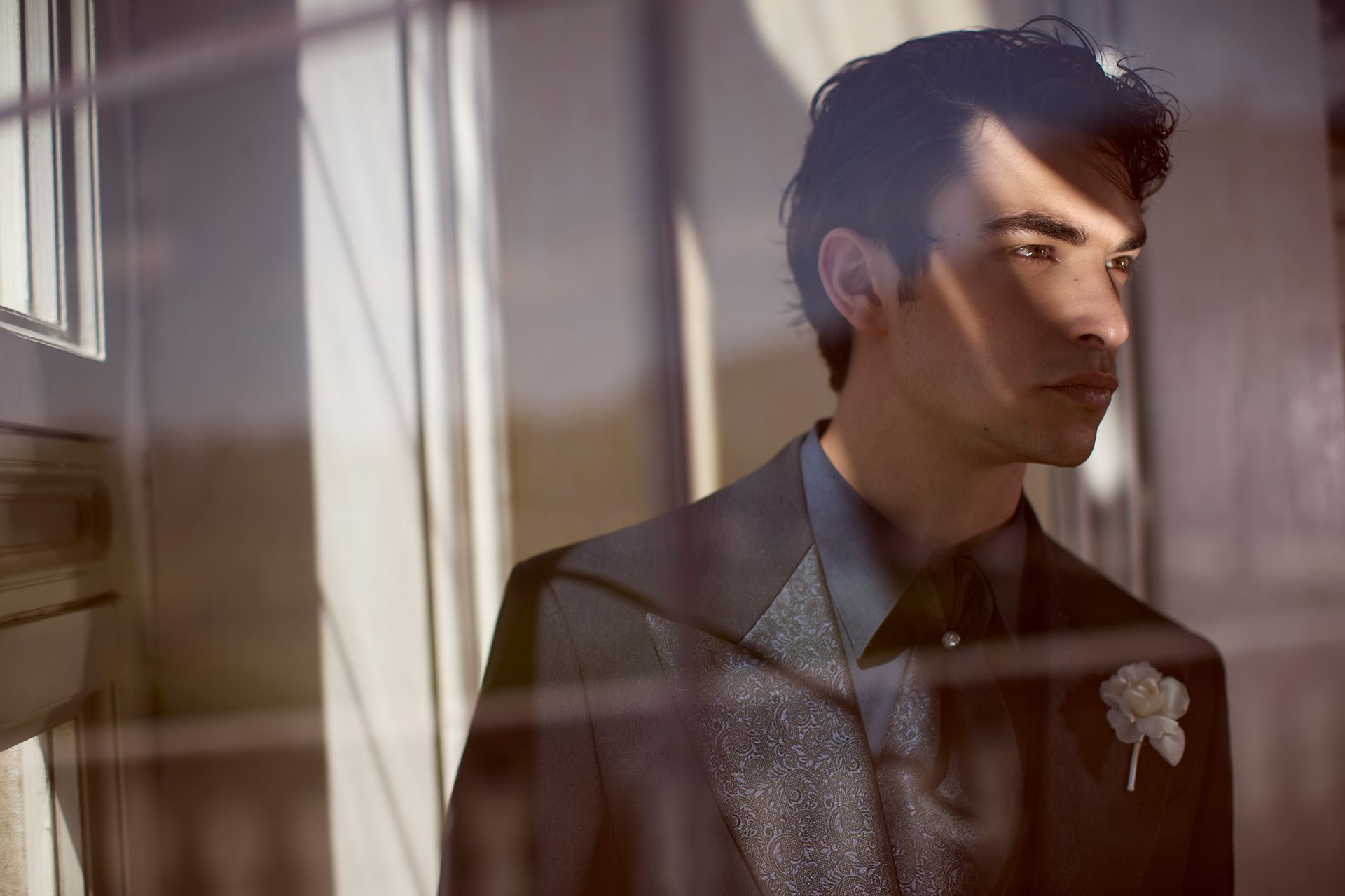 Uomo in giacca e cravatta che guarda di lato, illuminato dalla luce del sole che filtra da una finestra. 