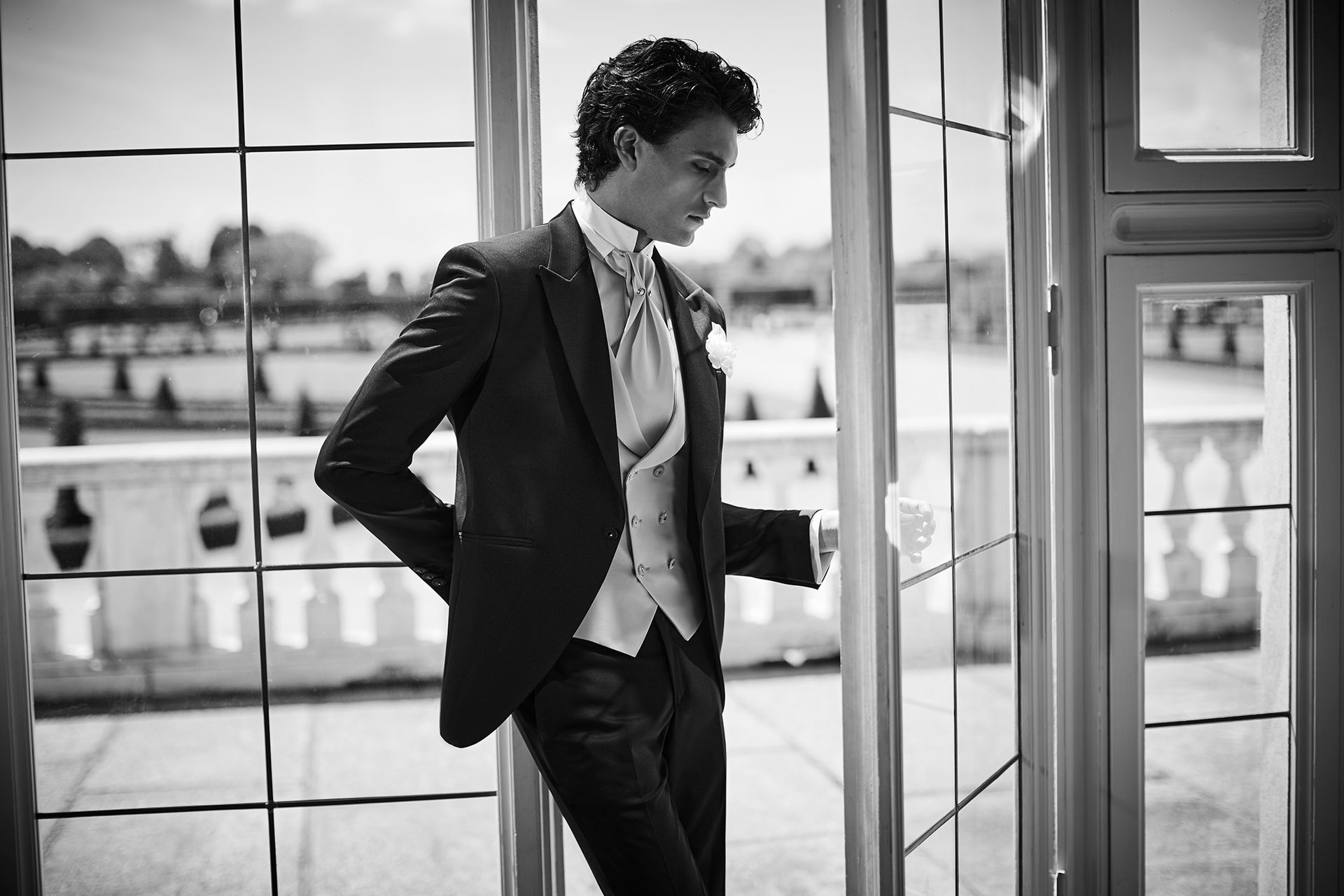 Uomo in giacca e cravatta che guarda in basso, appoggiato a una finestra con vista su un balcone e sull'ambiente esterno.