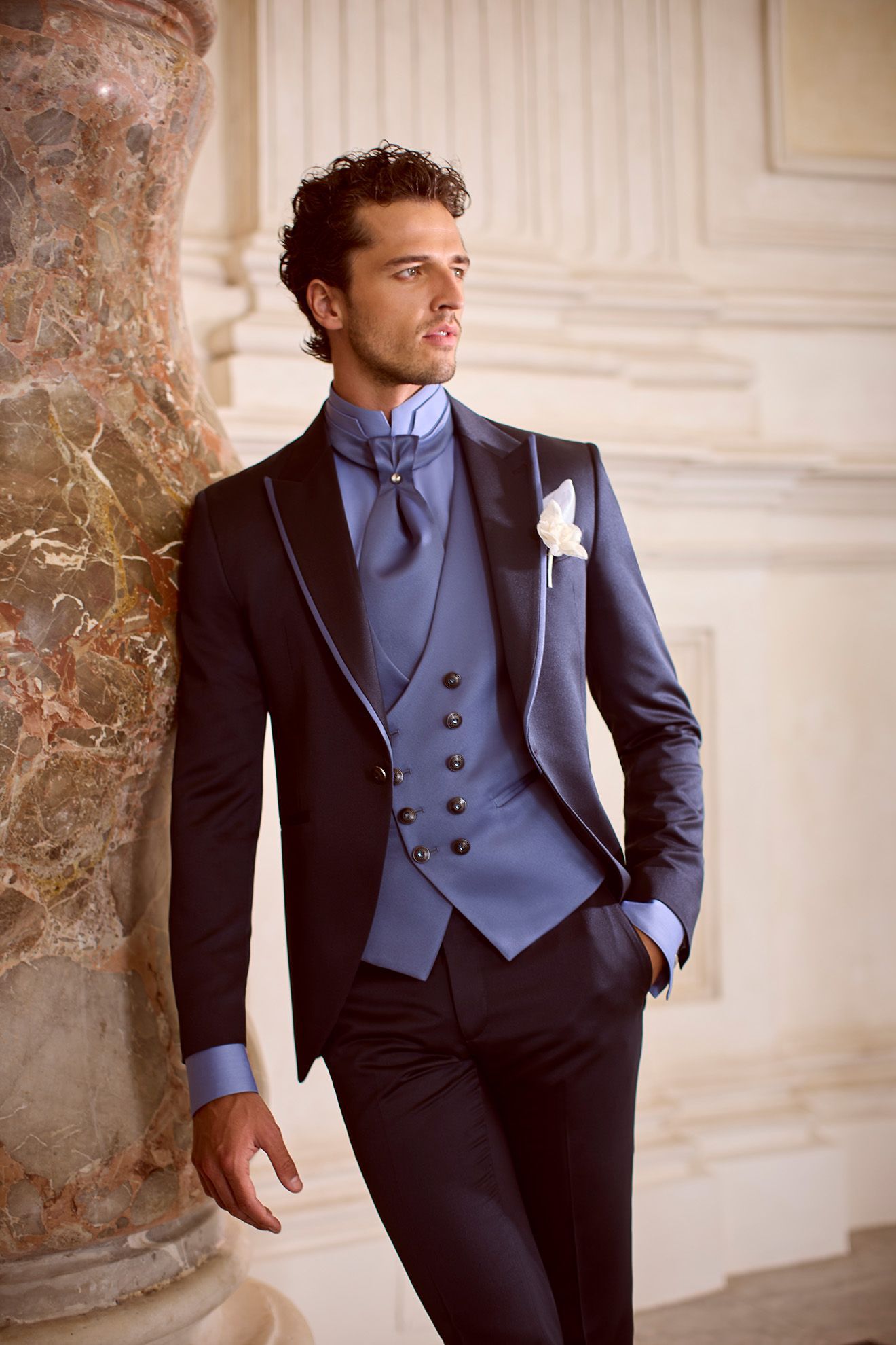 Uomo in abito blu, appoggiato a un pilastro di marmo, che guarda altrove.