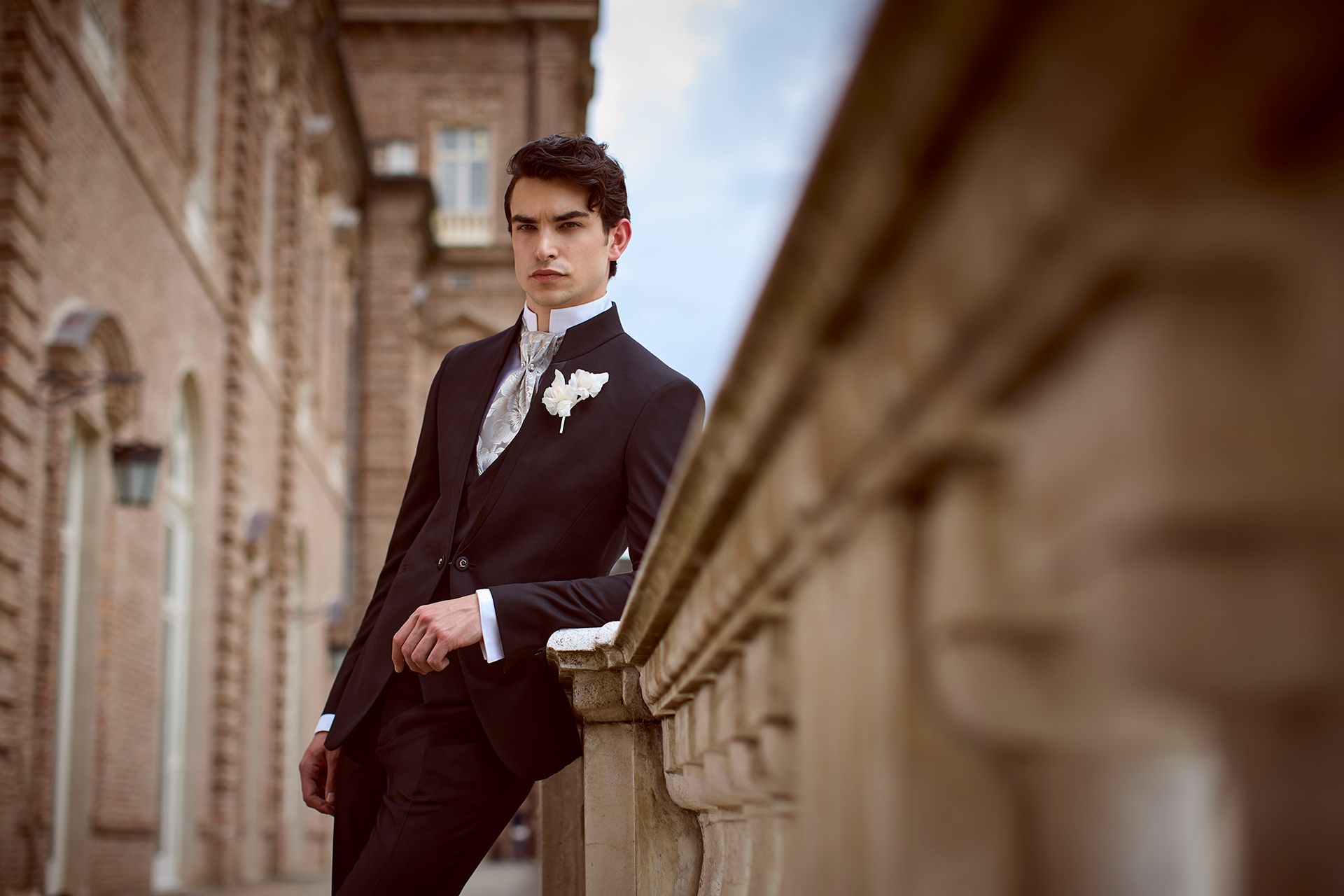 Uomo in abito nero formale appoggiato a una ringhiera di pietra all'aperto.