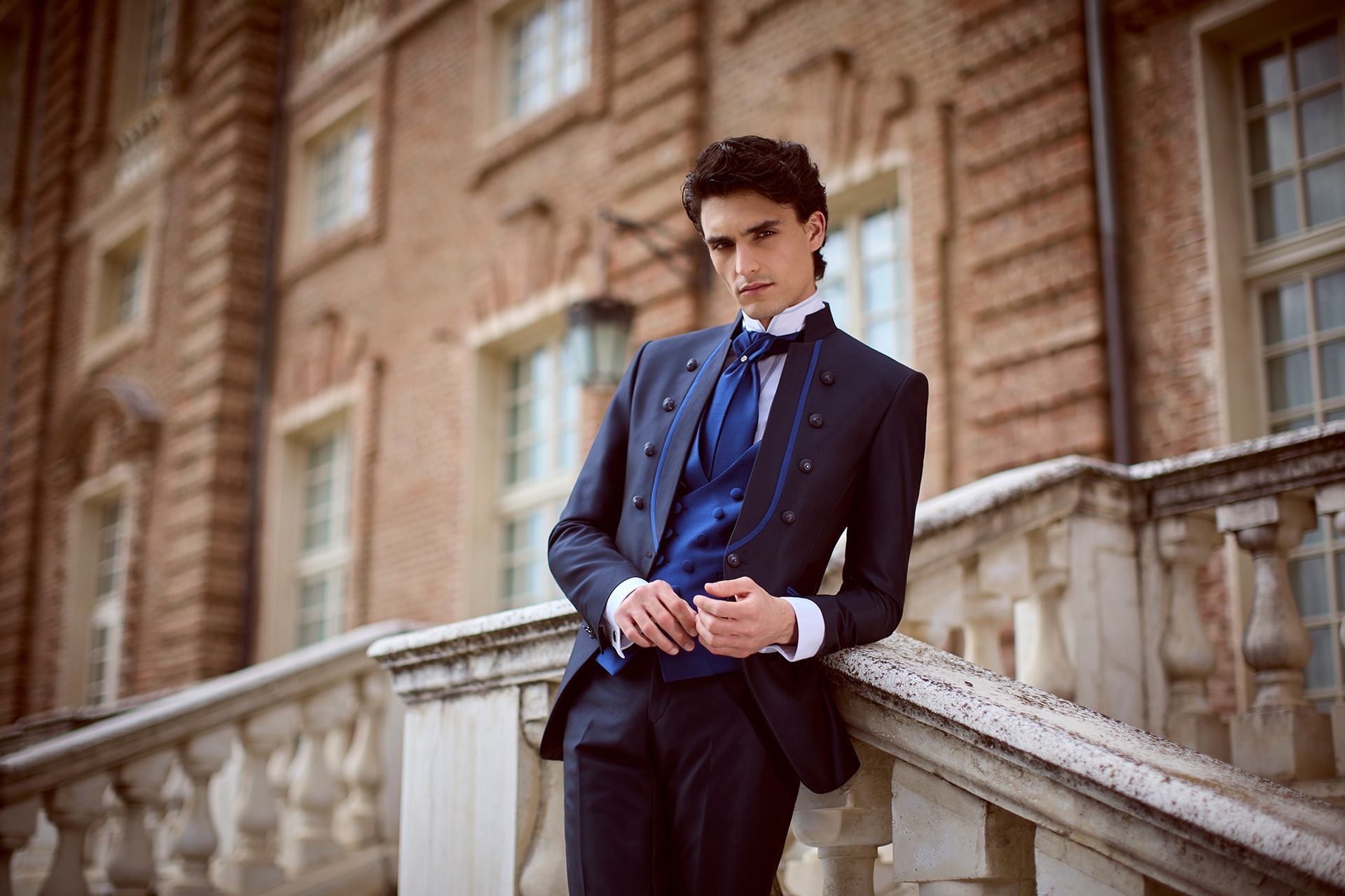 Uomo in abito blu navy, appoggiato a una balaustra in pietra. Gilet blu, polsini bianchi e ambiente formale.
