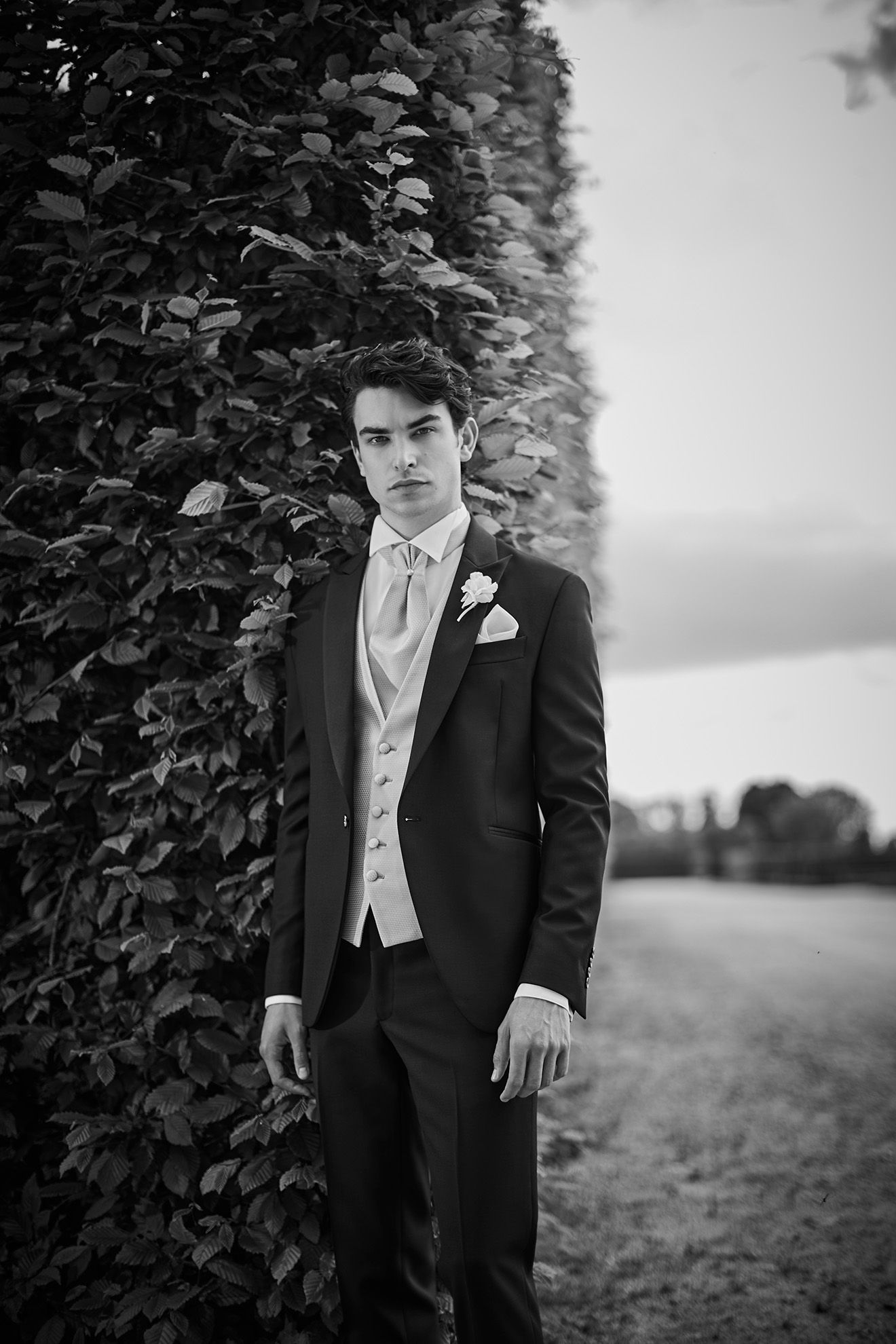 Un uomo in smoking è in piedi vicino a una siepe e guarda avanti. Foto in bianco e nero.