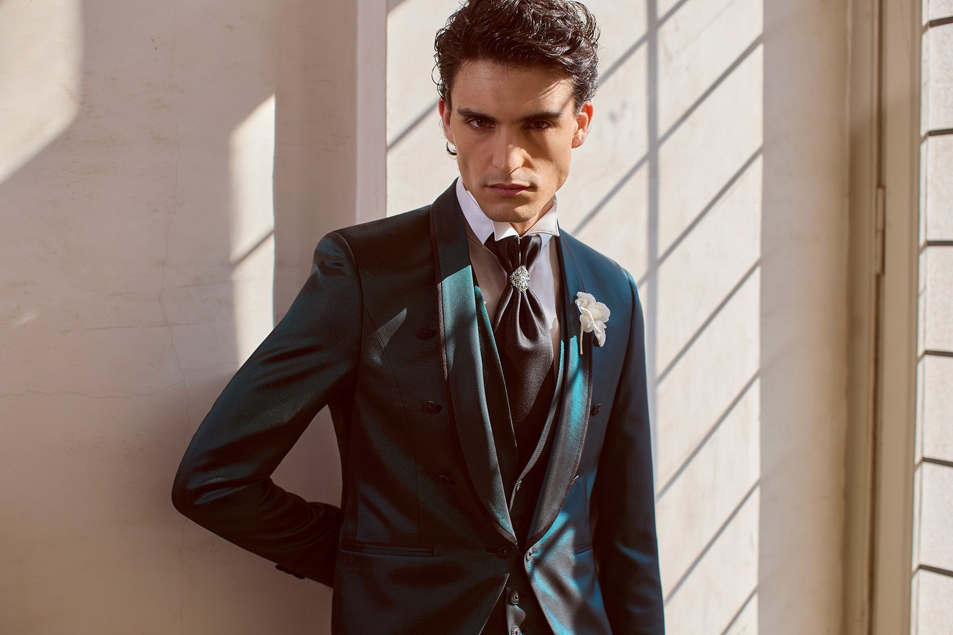 Uomo in smoking verde acqua, appoggiato a un muro vicino a una finestra, che guarda direttamente l'osservatore.