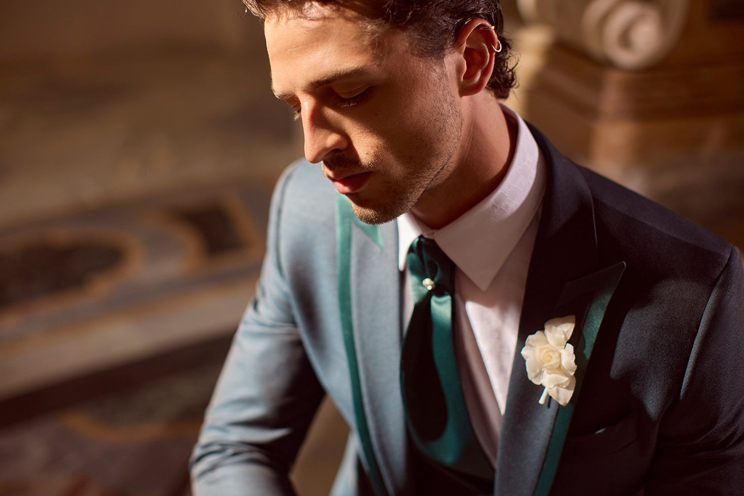 Uomo in abito verde acqua e cravatta, camicia bianca, sguardo basso, con fiore all'occhiello.