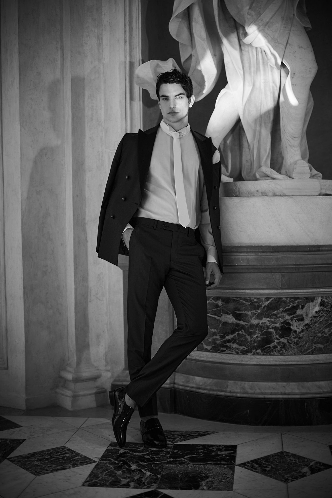 Un uomo in giacca e cravatta posa in una stanza con una statua e un pavimento a scacchi. Giacca sulla spalla, appoggiato.