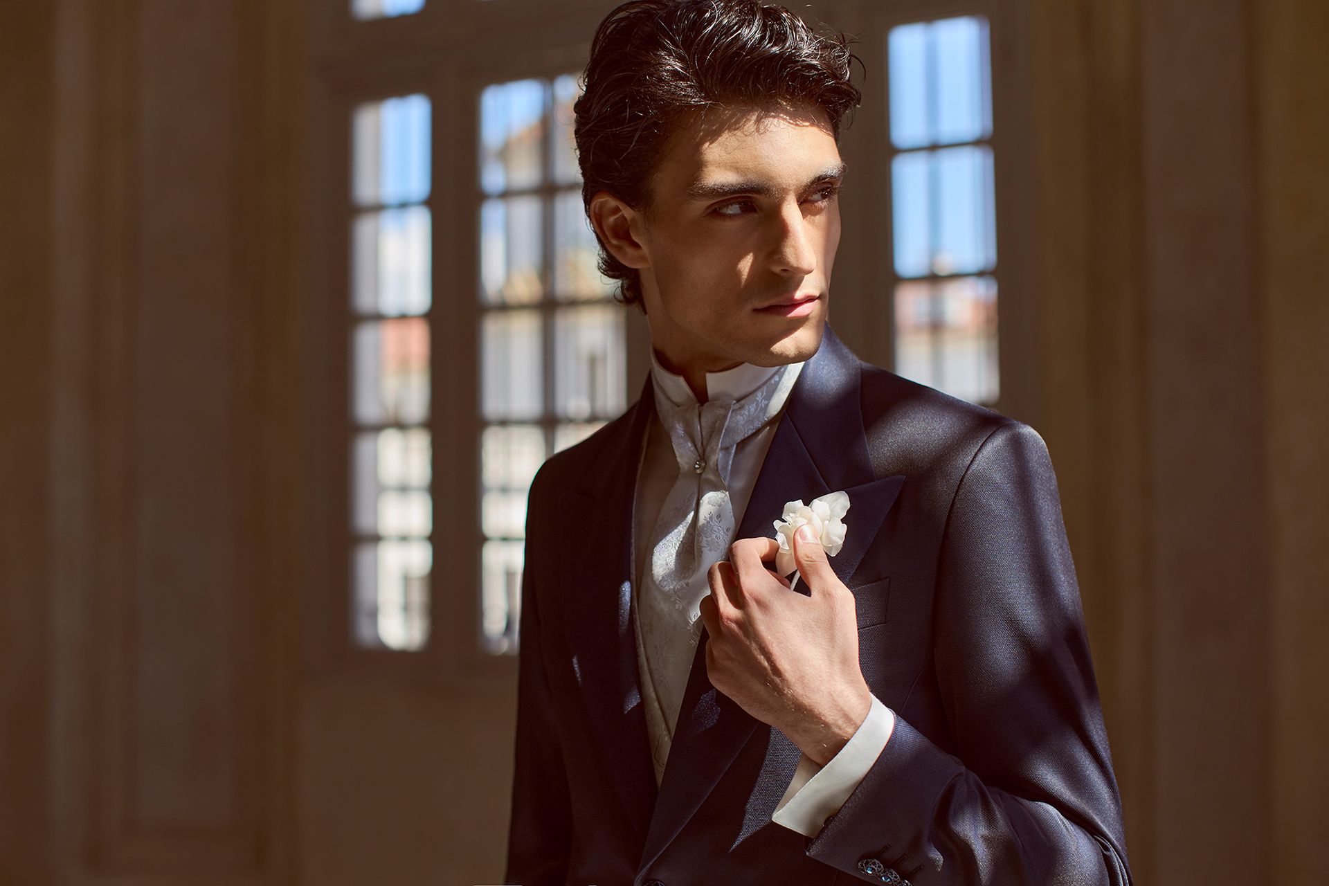 Uomo in abito scuro con fazzoletto da taschino bianco, in piedi vicino a una finestra illuminata dal sole.