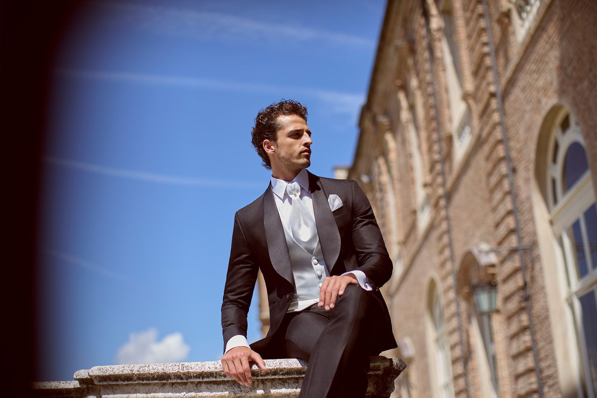 Uomo in abito formale, seduto su una struttura in pietra, che guarda lontano. Giornata di sole, edificio sullo sfondo.