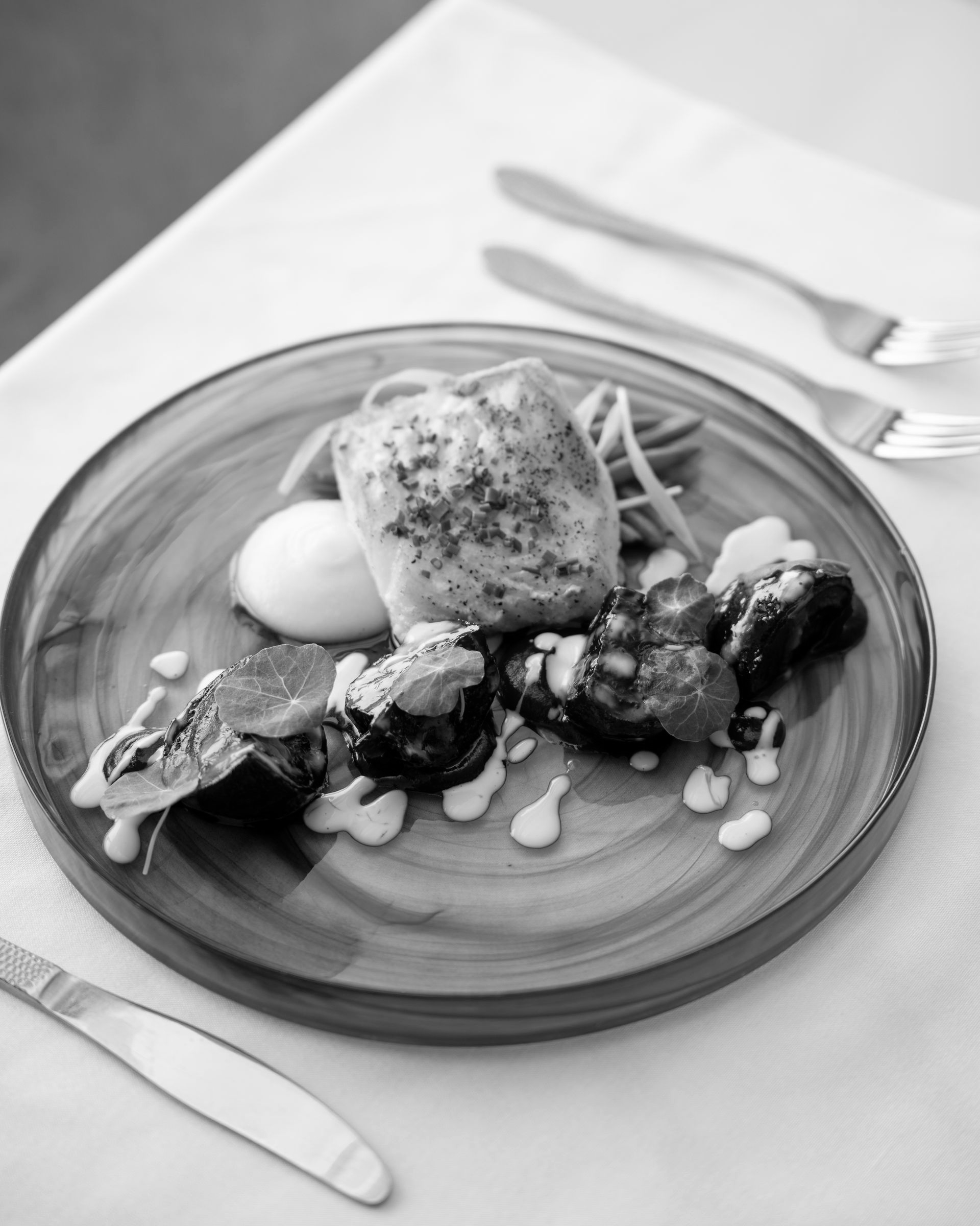 Fish and vegetables on a plate with white sauce, cutlery on the side, and a white tablecloth.